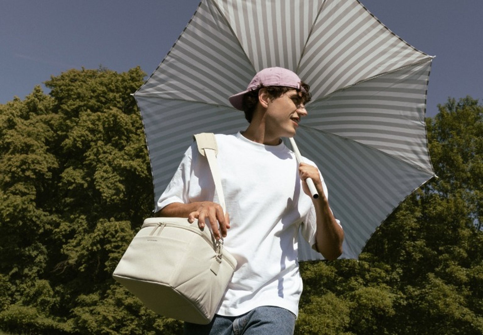junger mann trägt im park einen sonnenschirm und eine beige picknicktasche von Kapten & Son