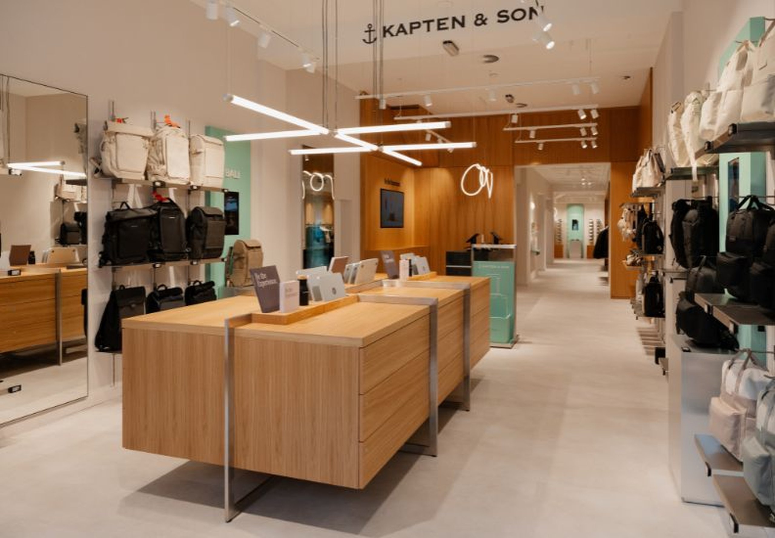 Un elegante interior de tienda con un perchero de chaquetas en tonos neutros y una pared con filas de gafas de sol. El letrero de Kapten & Son está iluminado al fondo.
