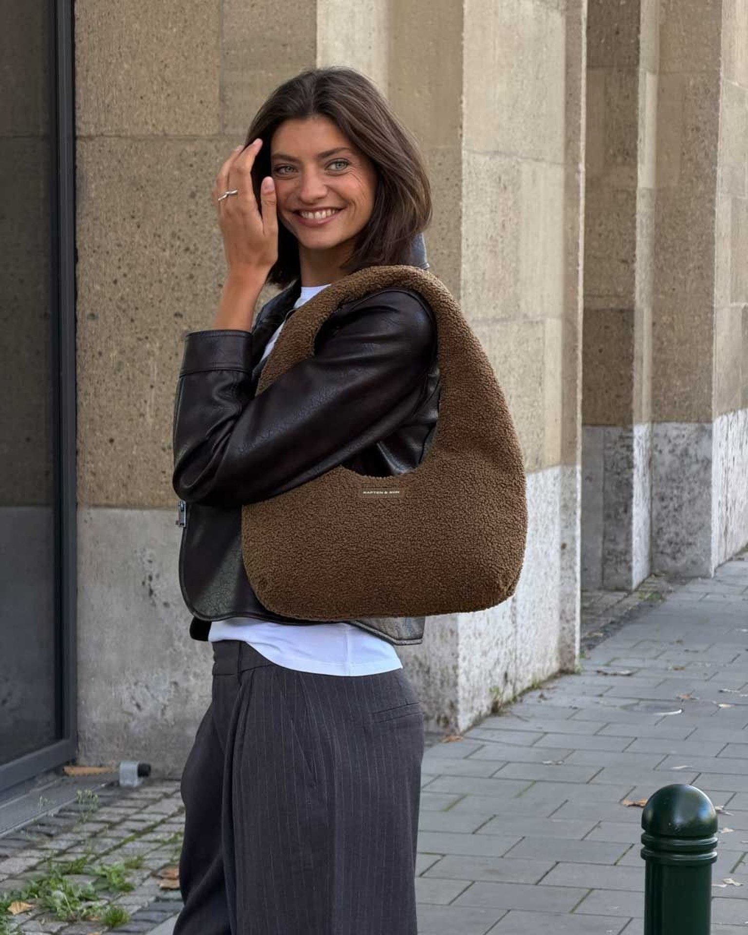 Lächelnde Frau mit einer Schultertasche aus Teddyplüsch auf einem Gehweg vor einem Steingebäude.
