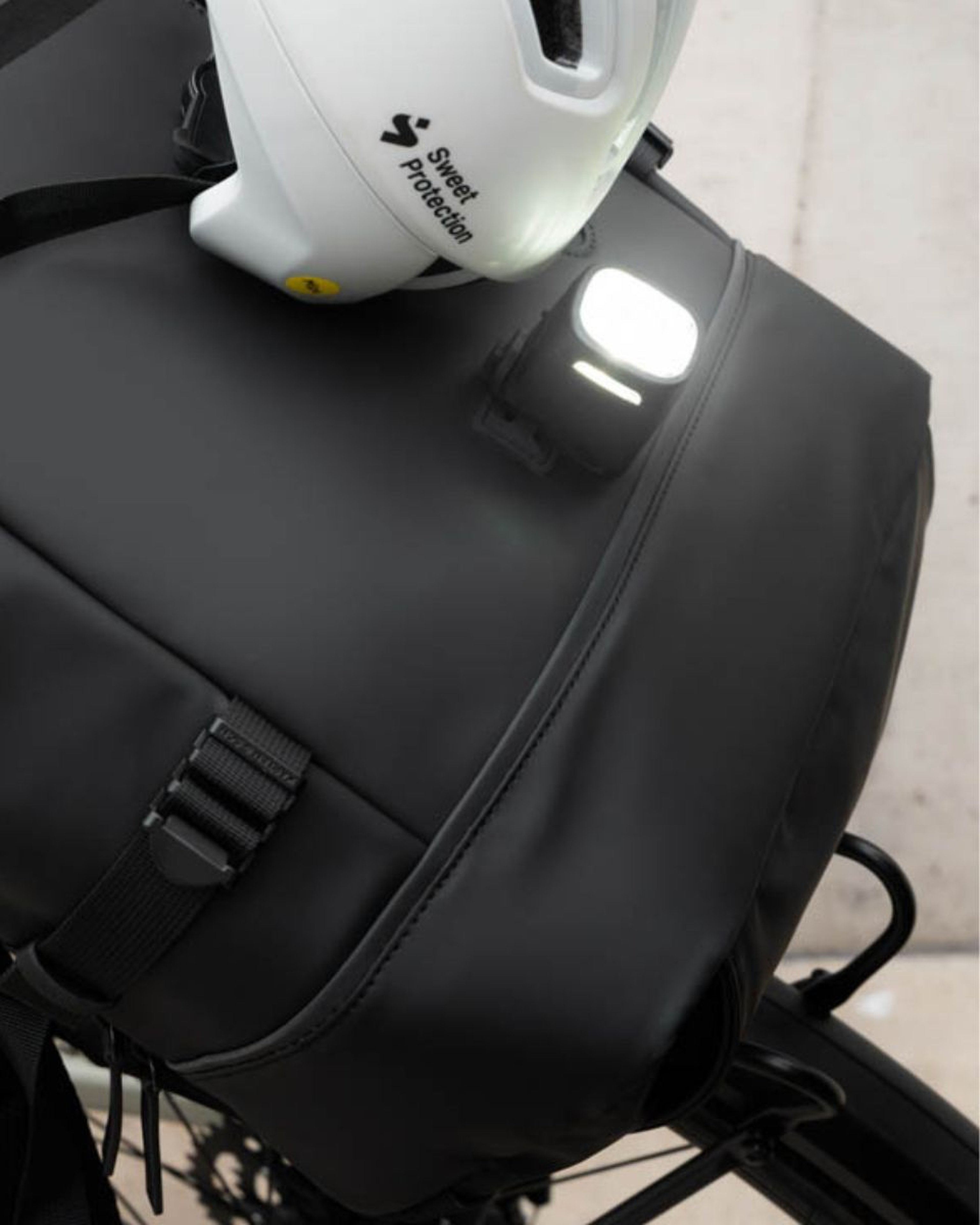Ein weißer Helm liegt auf einer schicken schwarzen Fahrradtasche mit einem angebrachten hellen, quadratischen Licht – ein modernes und sicheres Fahrrad-Setup.