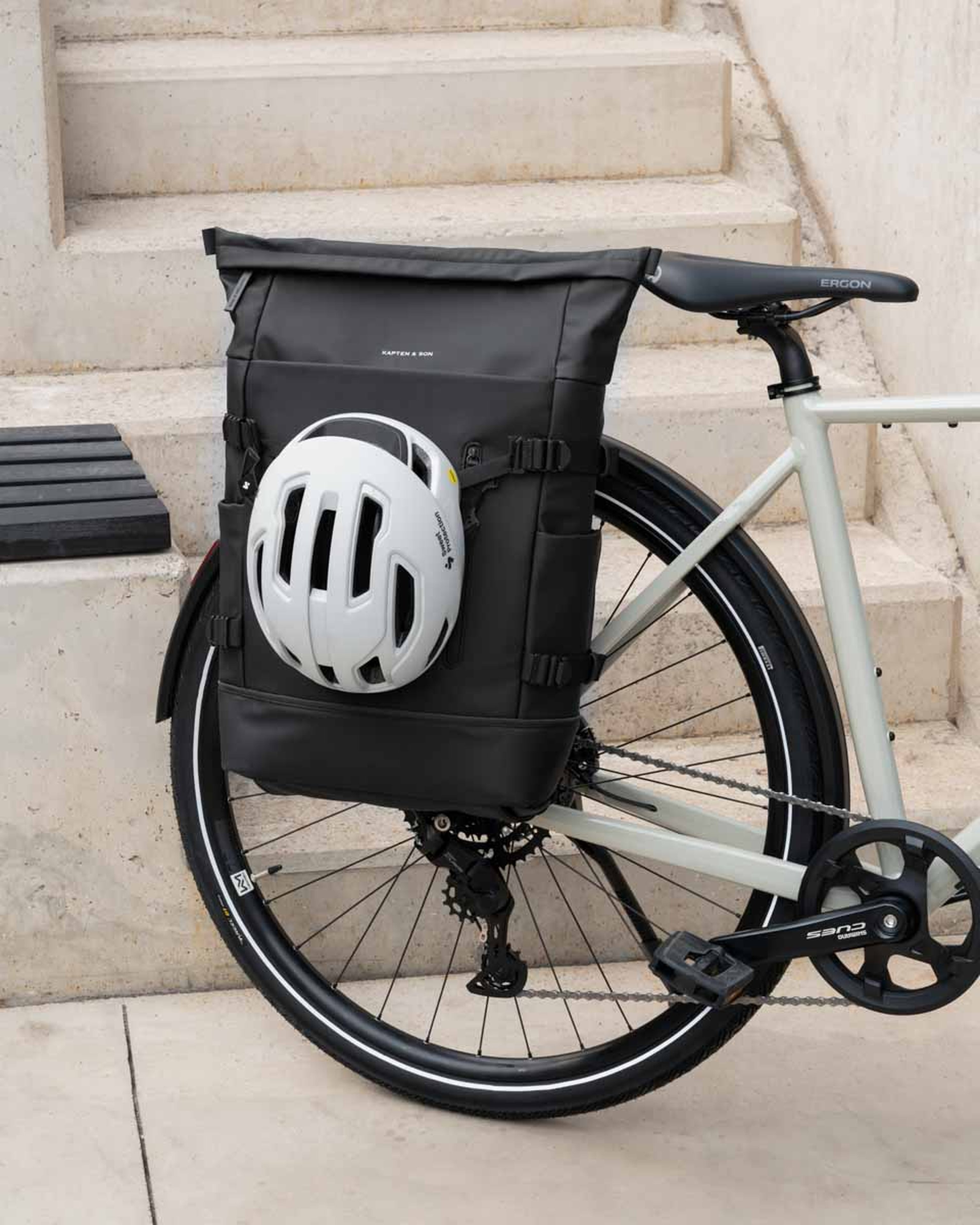 Ein schicker schwarzer Rucksack ist am hinteren Gepäckträger eines Fahrrads befestigt, mit einem daran geschnallten weißen Helm. Das Fahrrad lehnt an Betonstufen und vermittelt Aufbruchstimmung.