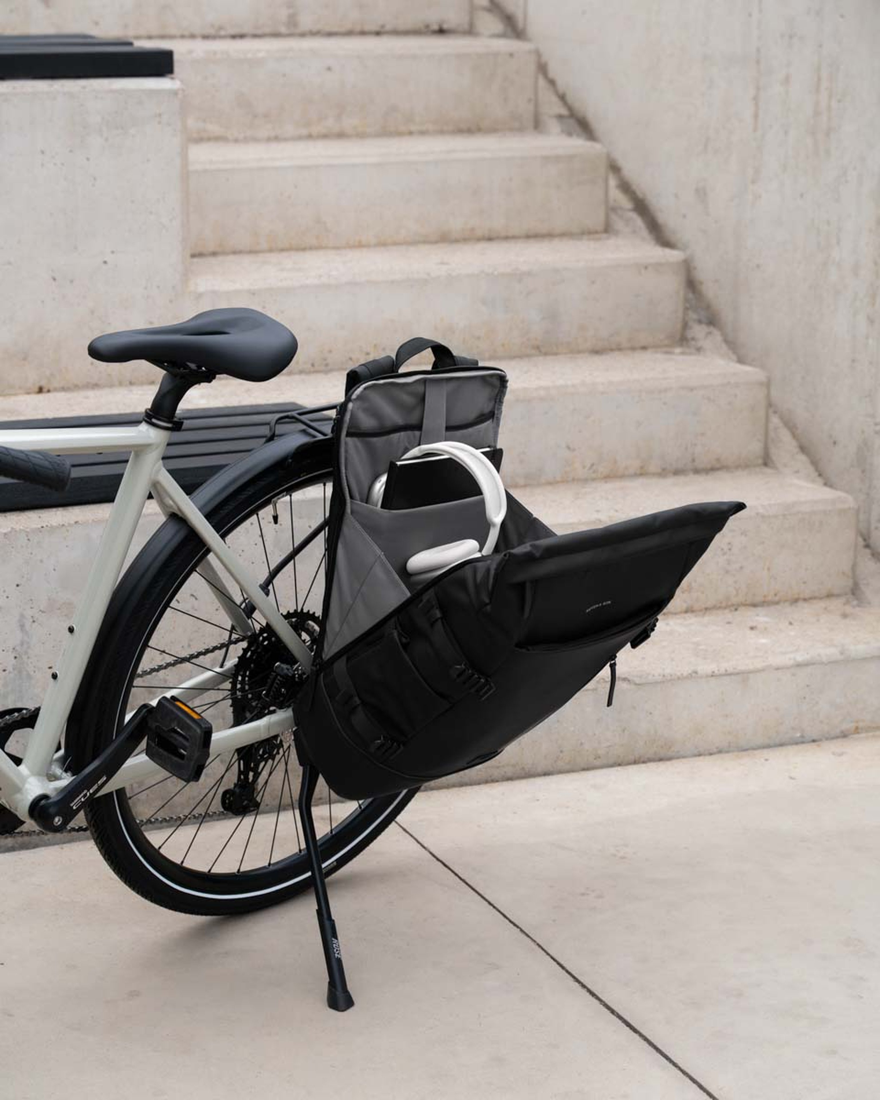 Ein geparktes Fahrrad mit einer geöffneten schwarzen Gepäcktasche am Heck, in der ein Laptop und weiße Kopfhörer zu sehen sind. Im Hintergrund sind Betonstufen zu erkennen.