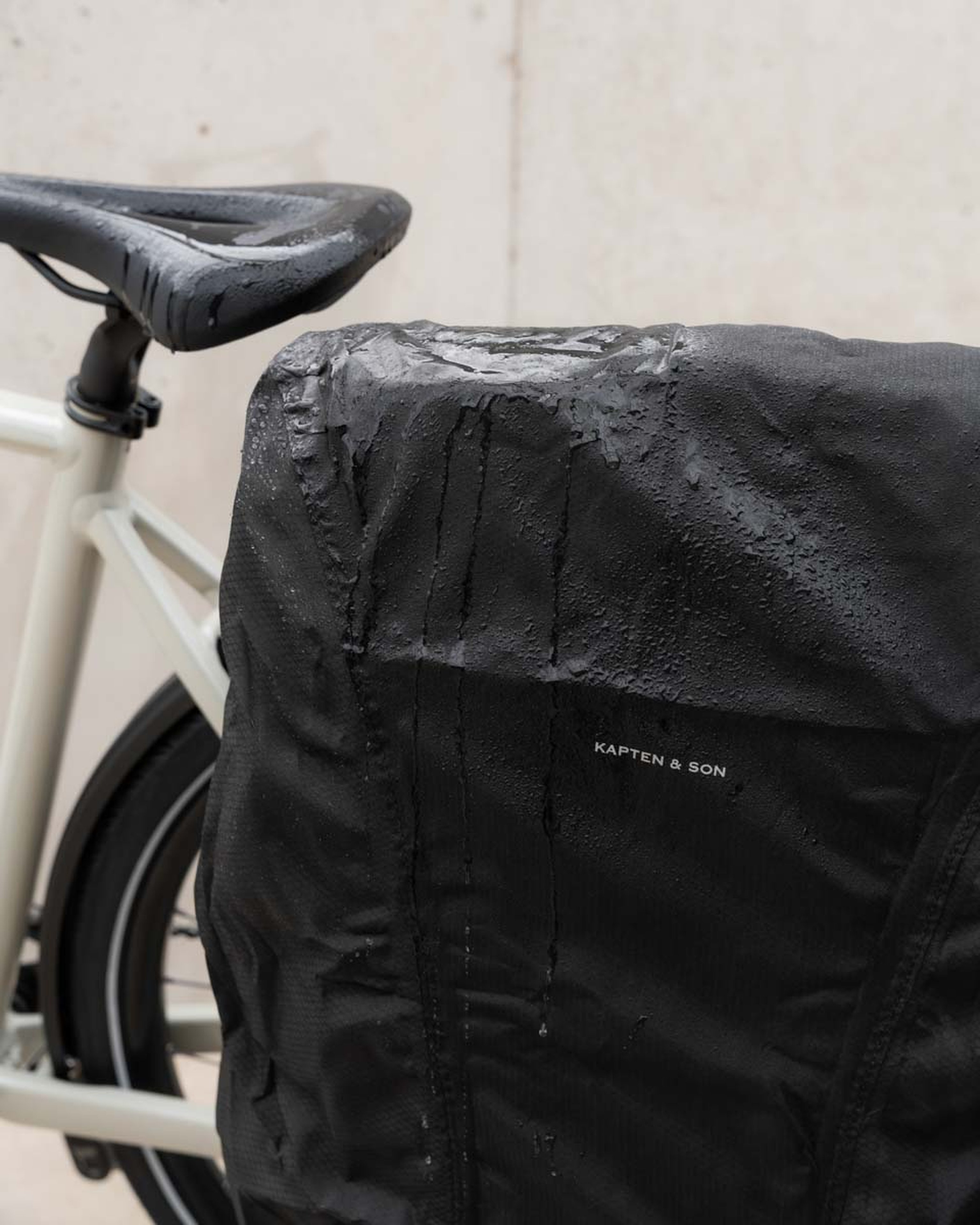 Ein regenbenetzter schwarzer Fahrradsattel und Rucksack mit sichtbaren Wassertropfen. Der Rucksack mit dem Aufdruck „Kapten & Son" befindet sich auf einem weißen Fahrrad und vermittelt eine Regentag-Atmosphäre.