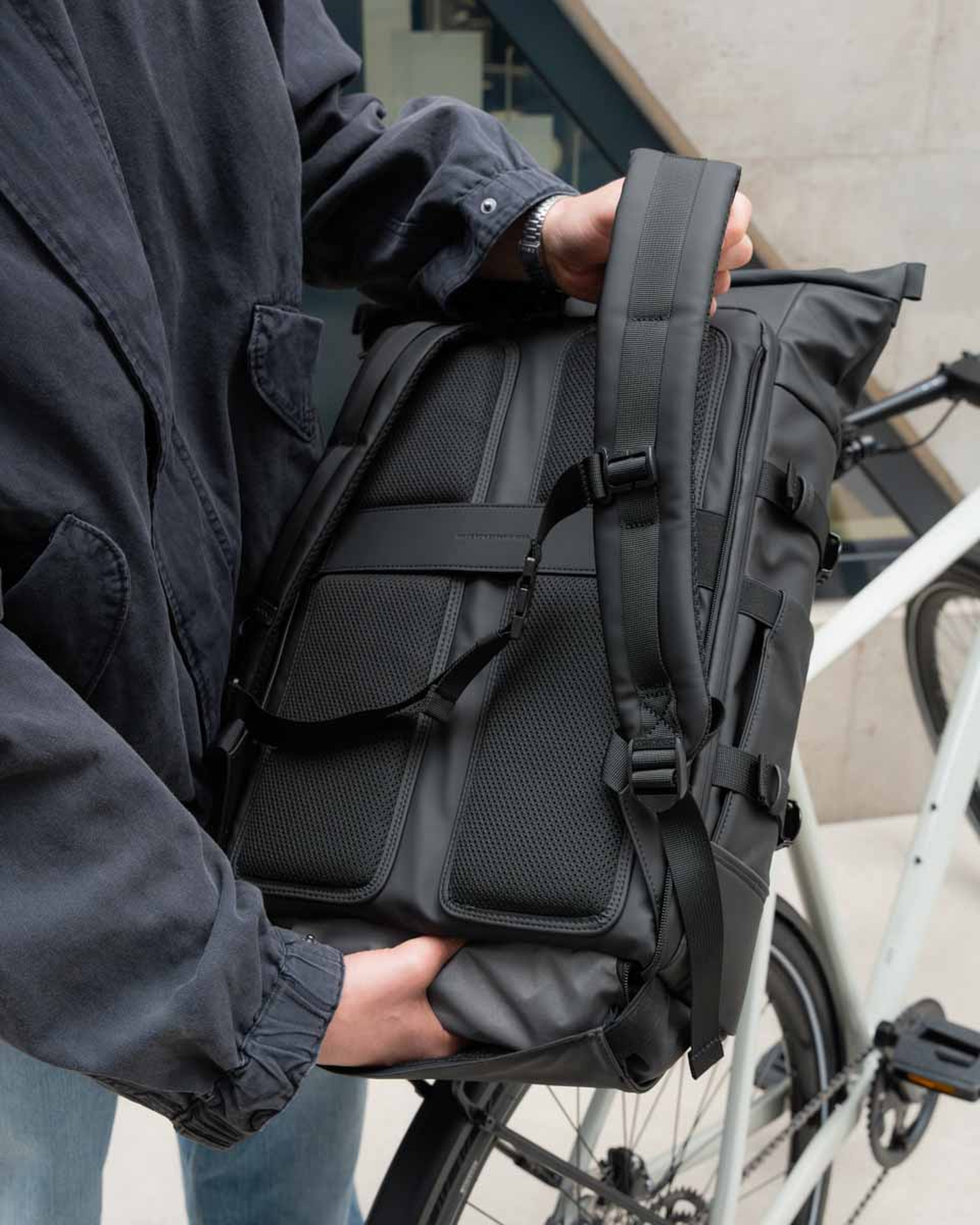 Eine Person in einer dunklen Jacke hält einen schicken schwarzen Rucksack mit gepolsterten Trägern neben einem weißen Fahrrad. Das Setting wirkt urban und modern.