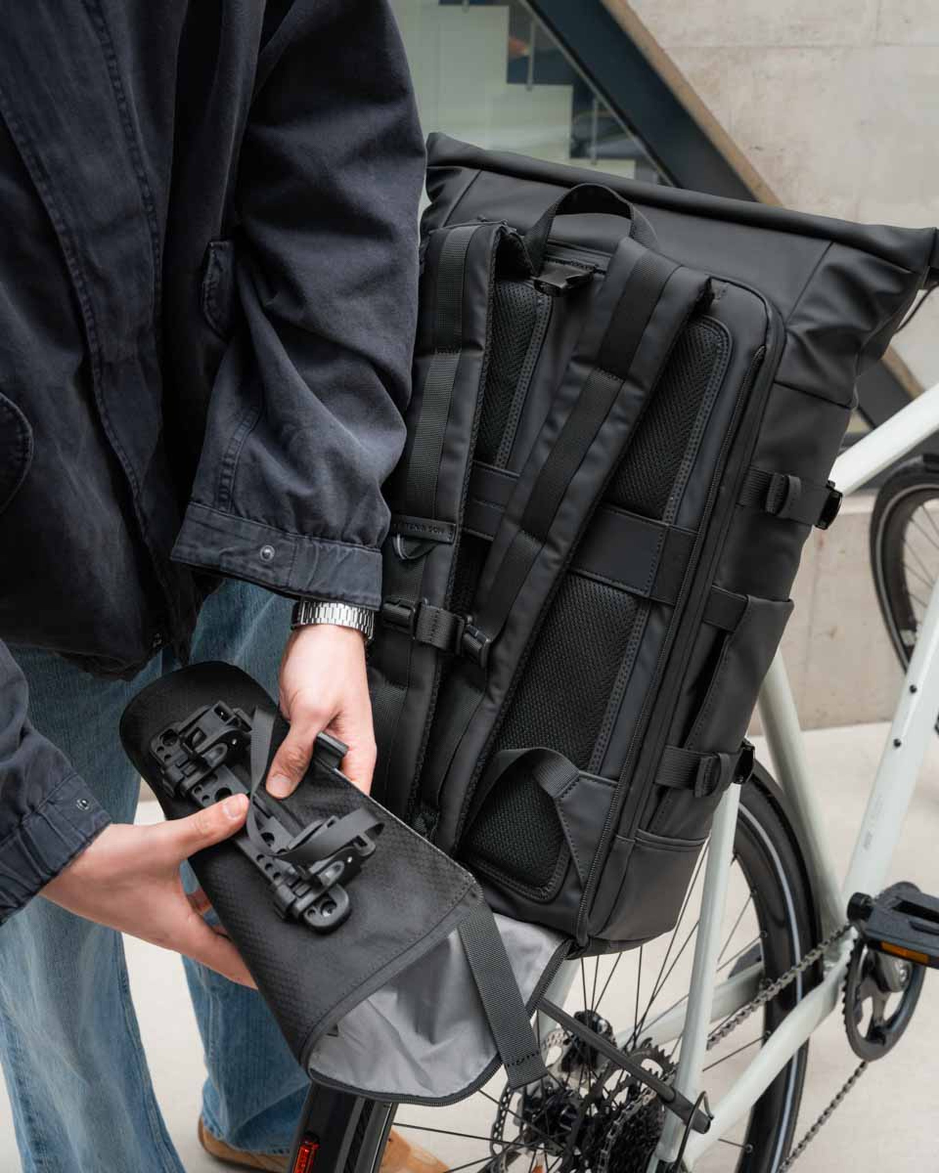 Eine Person in einer dunklen Jacke befestigt einen schwarzen, robusten Rucksack an einem Fahrrad. Die Szene vermittelt Praktikabilität und Aufbruchstimmung für den täglichen Arbeitsweg.