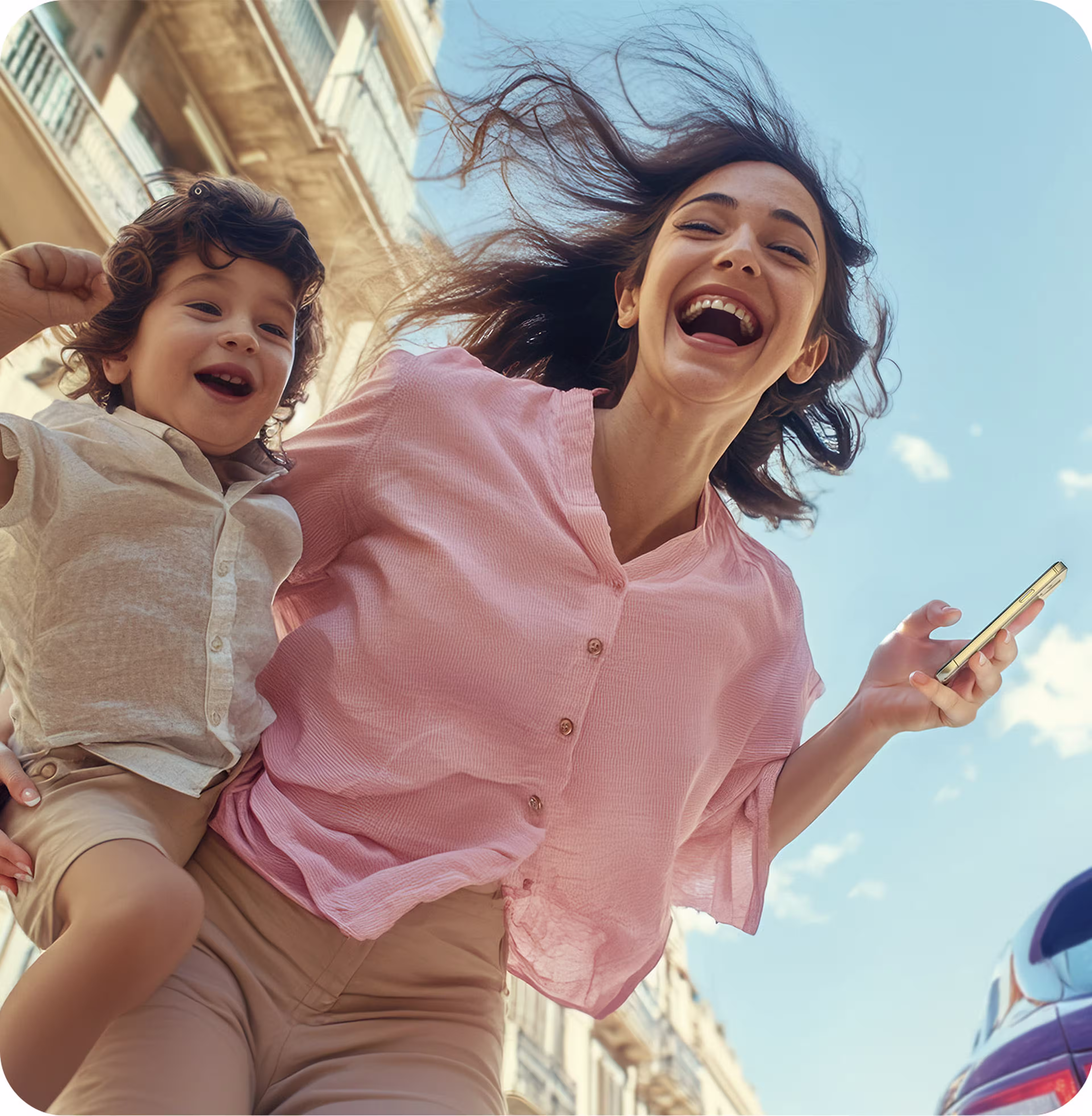 A joyful woman and child, both smiling, outdoors. The woman holds a phone, and the sky and buildings are visible in the background.