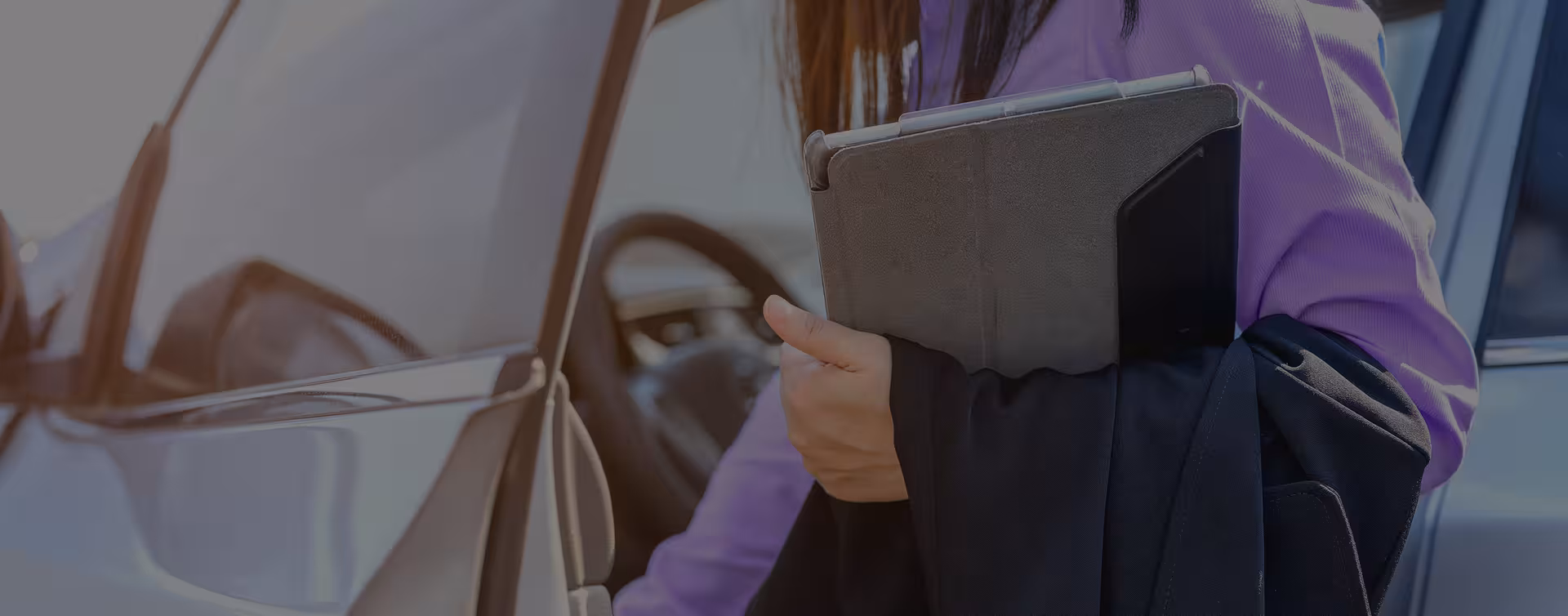 A person standing near a car door holding a grey tablet in one hand, wearing a purple top.