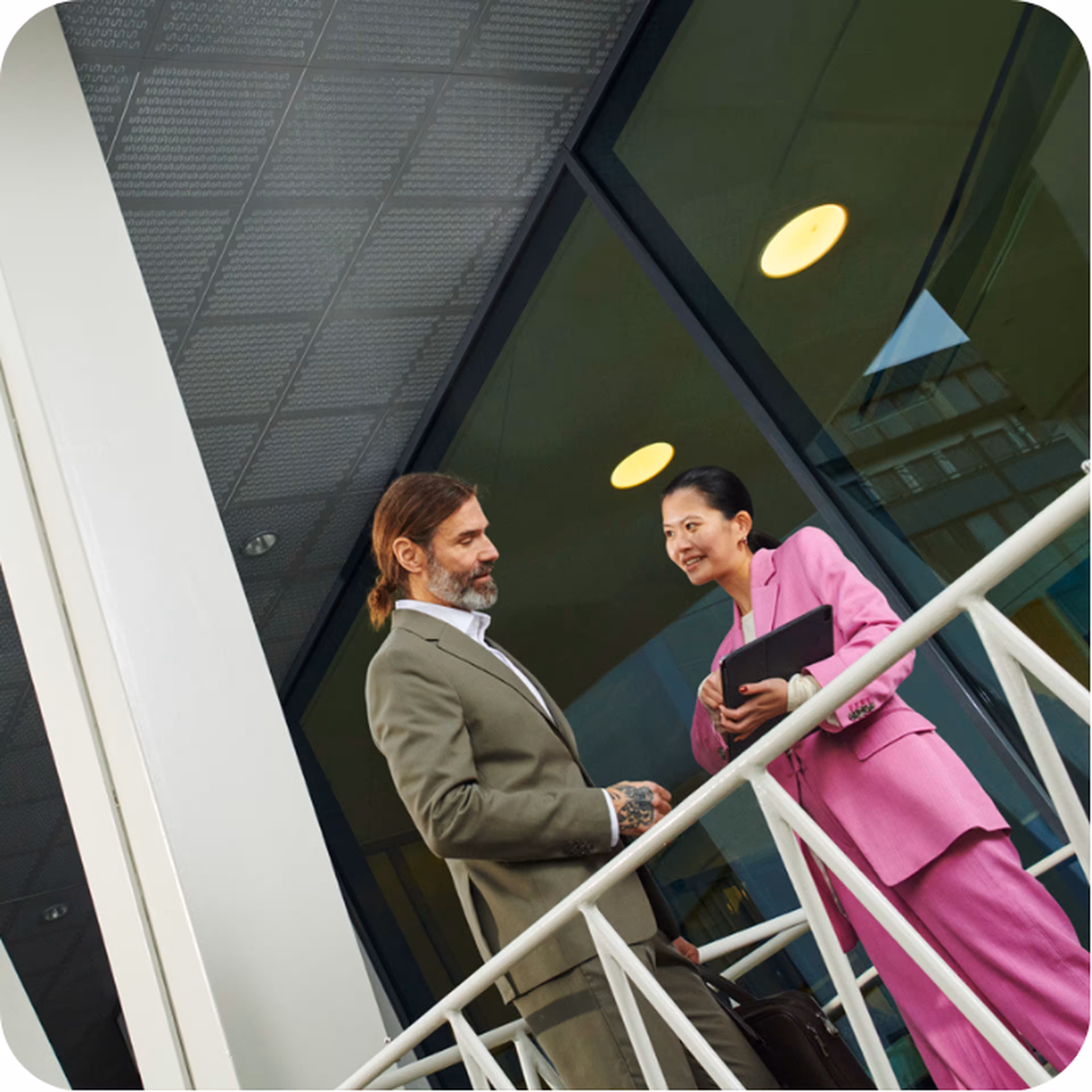 Two business professionals, a man and a woman, conversing outdoors near a modern building with large windows and railing.
