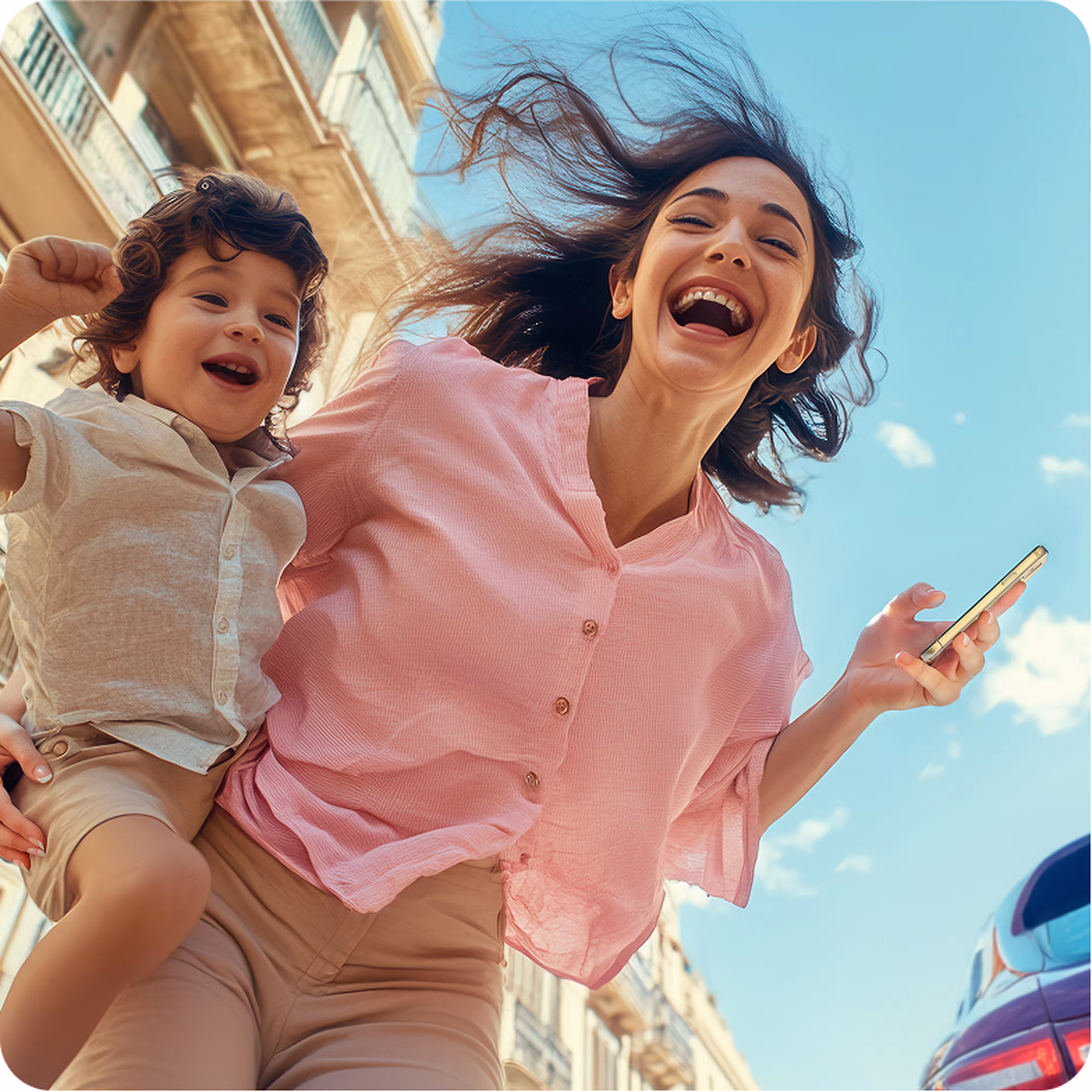 A joyful woman holding a phone and a smiling child outdoors, both laughing with wind-blown hair against a clear blue sky.
