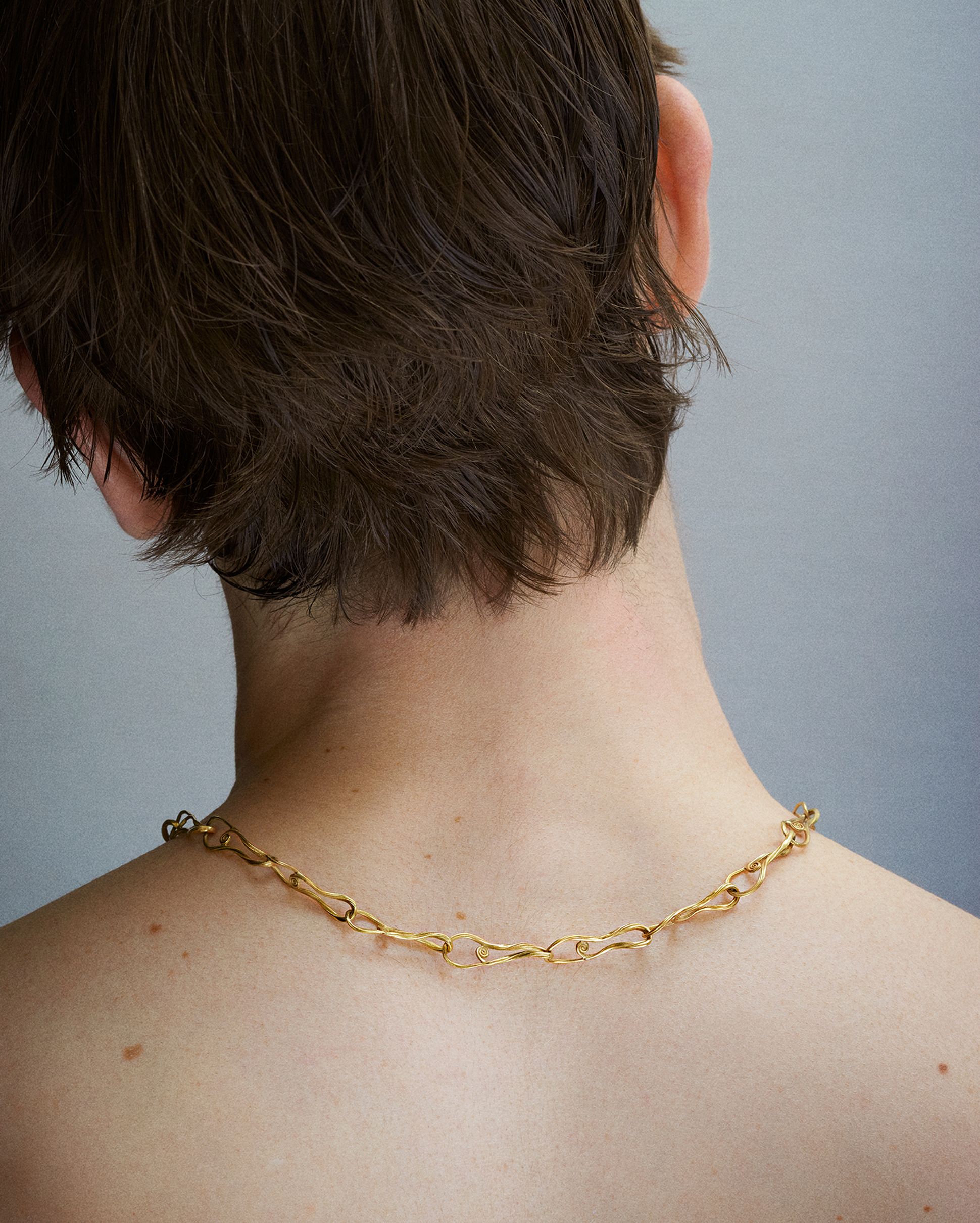 Back view of a person wearing a sculptural gold chain necklace by Griegst, resting on bare shoulders, with short tousled hair and a soft grey background.