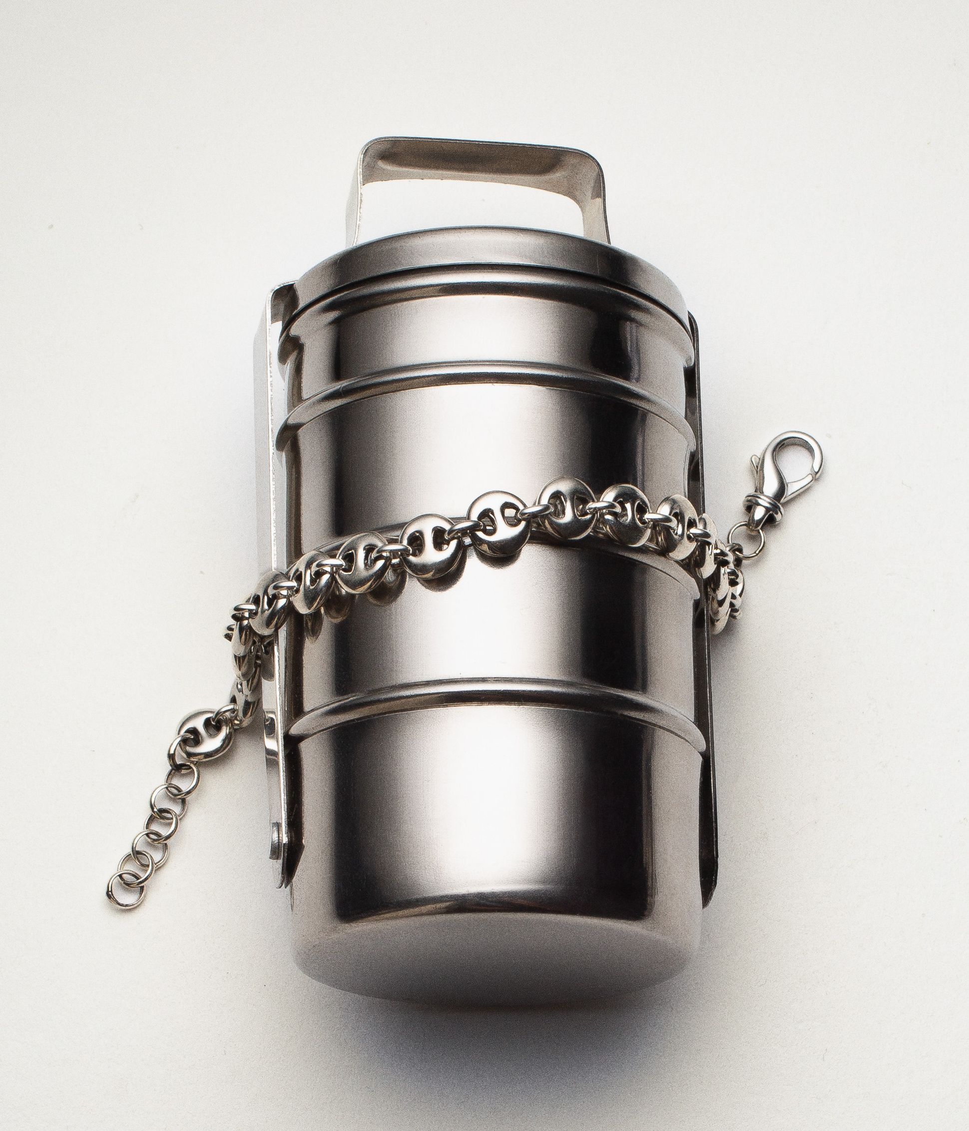 Petit objet cylindrique en métal argenté, ressemblant à une boîte ou un étui hermétique, orné d’une chaîne argentée avec fermoir mousqueton enroulée autour.