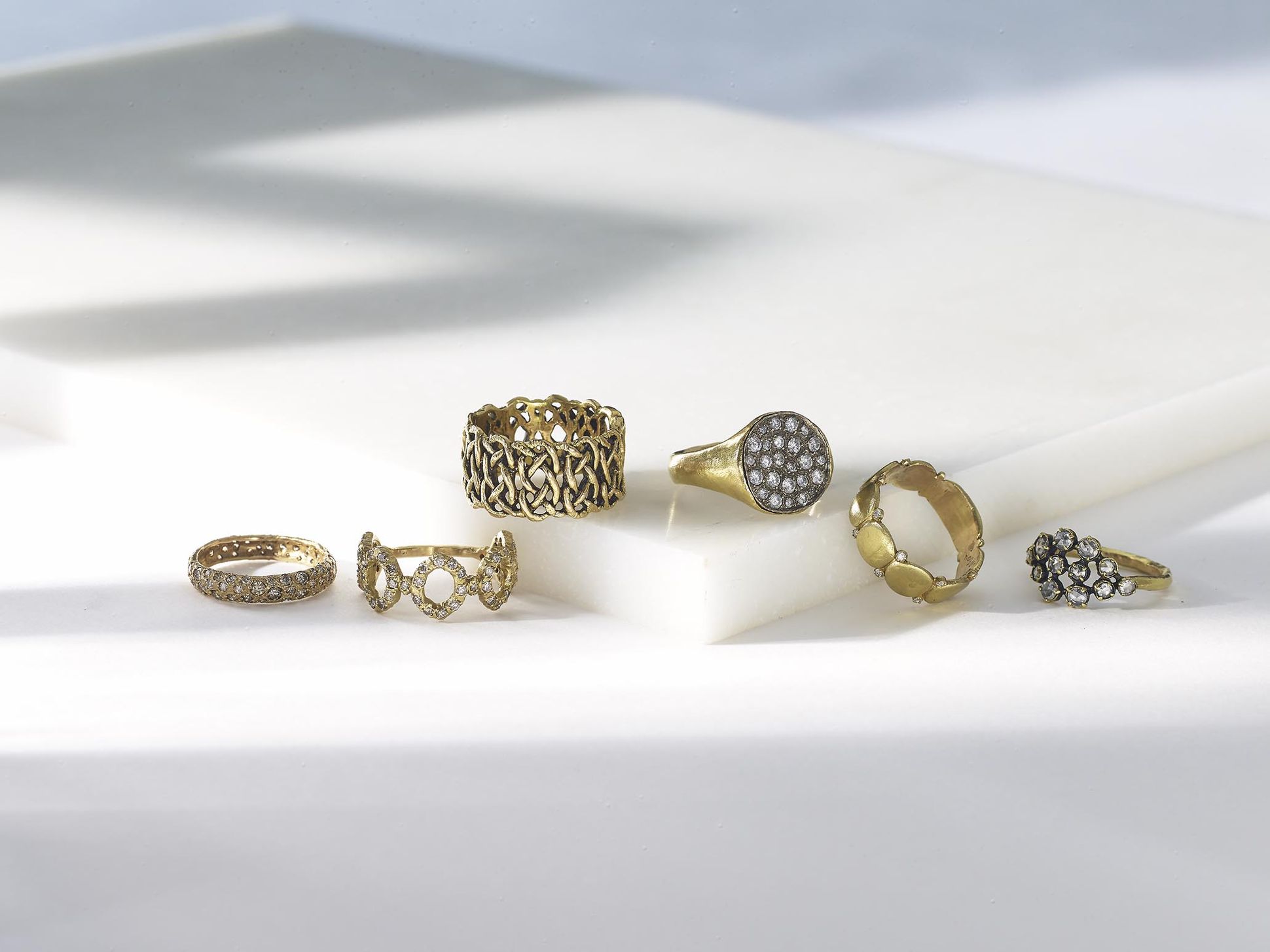Sélection de bagues en or jaune et diamants, aux designs variés, présentées sur un support en marbre blanc sous une lumière naturelle.