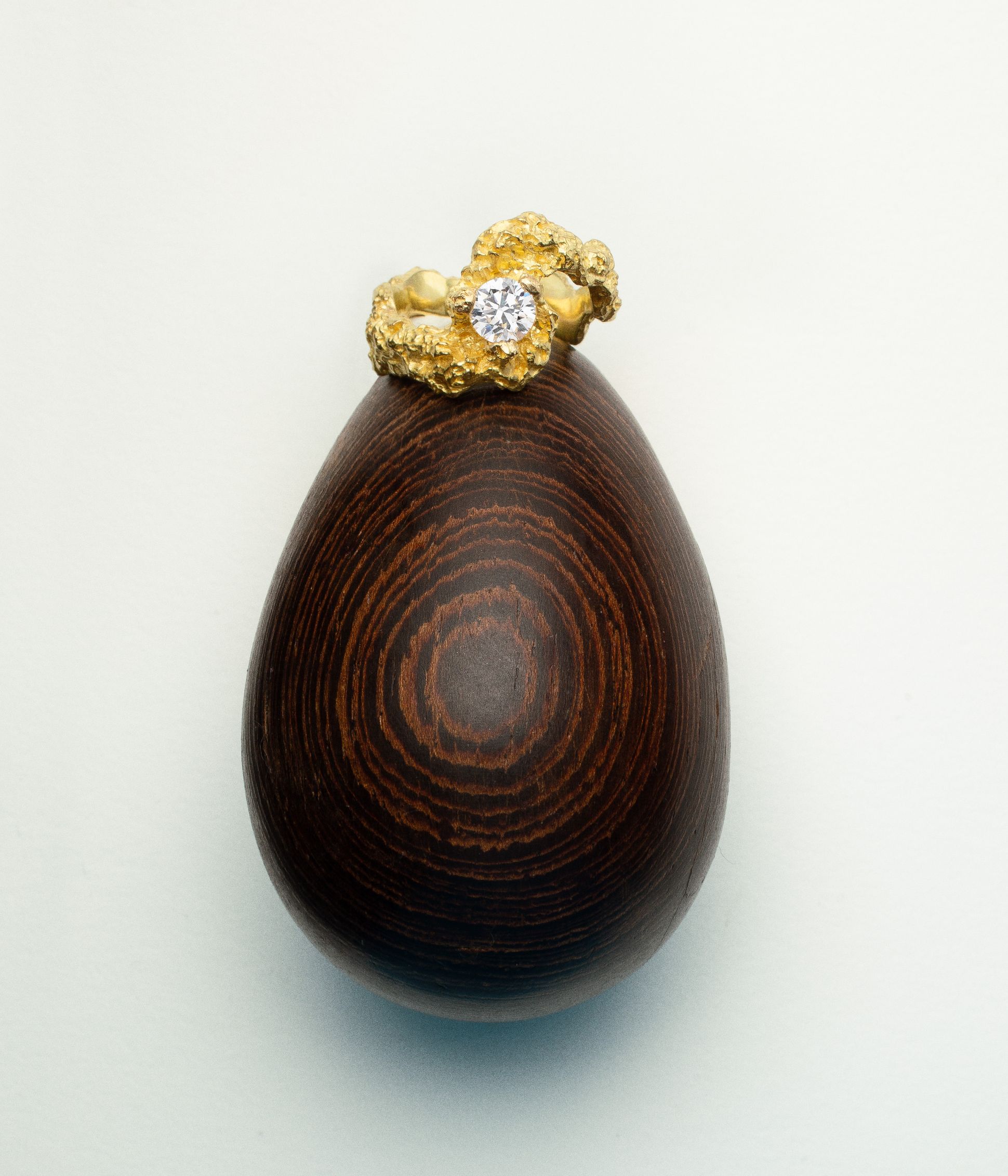 Bague sculpturale en or jaune texturé sertie d’un diamant rond, posée sur un galet en bois sombre à la texture veinée.