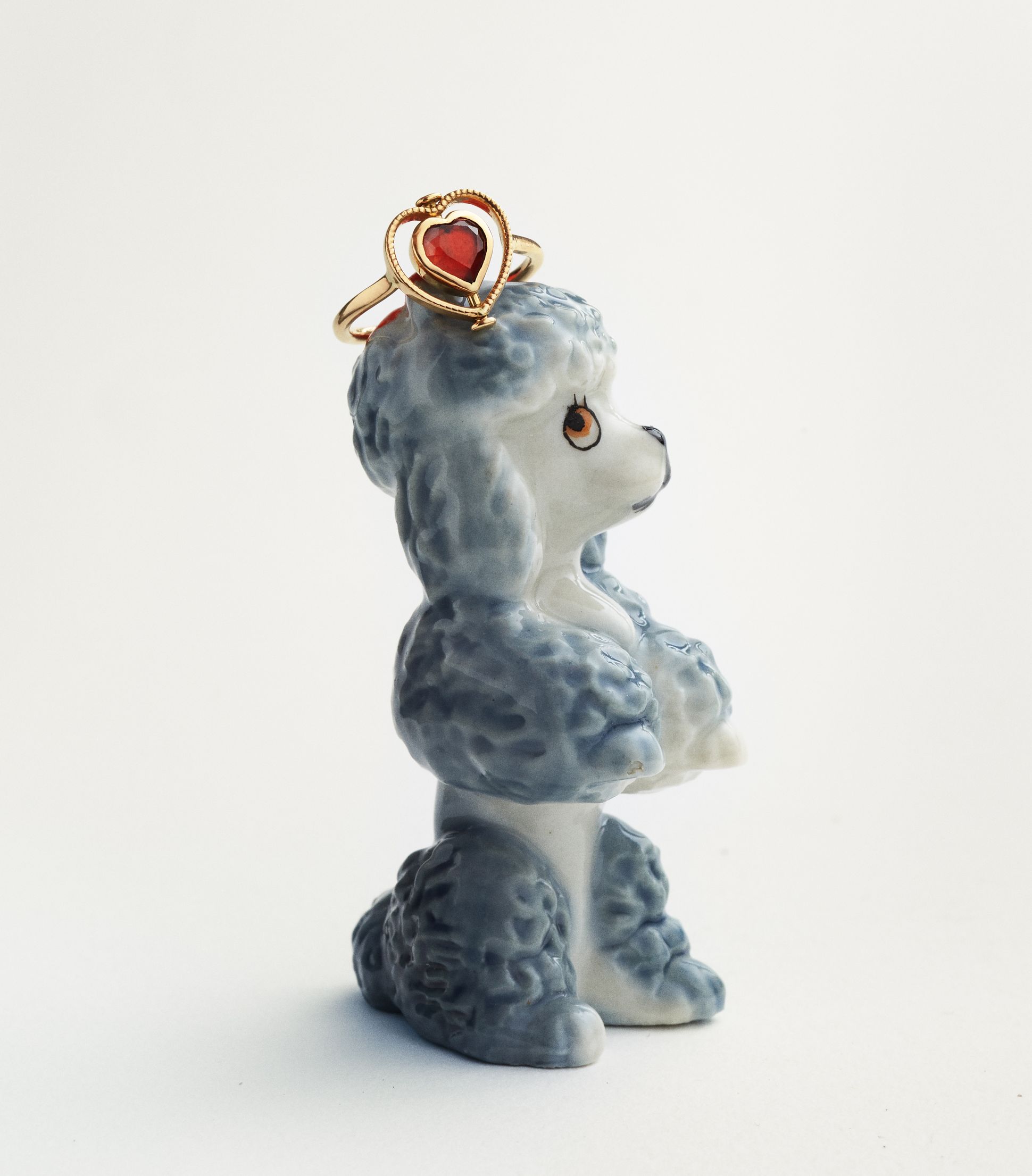 Ceramic poodle figurine used as a display, with a Dorette gold ring featuring a red heart-shaped gemstone placed on its head, photographed on a light background.