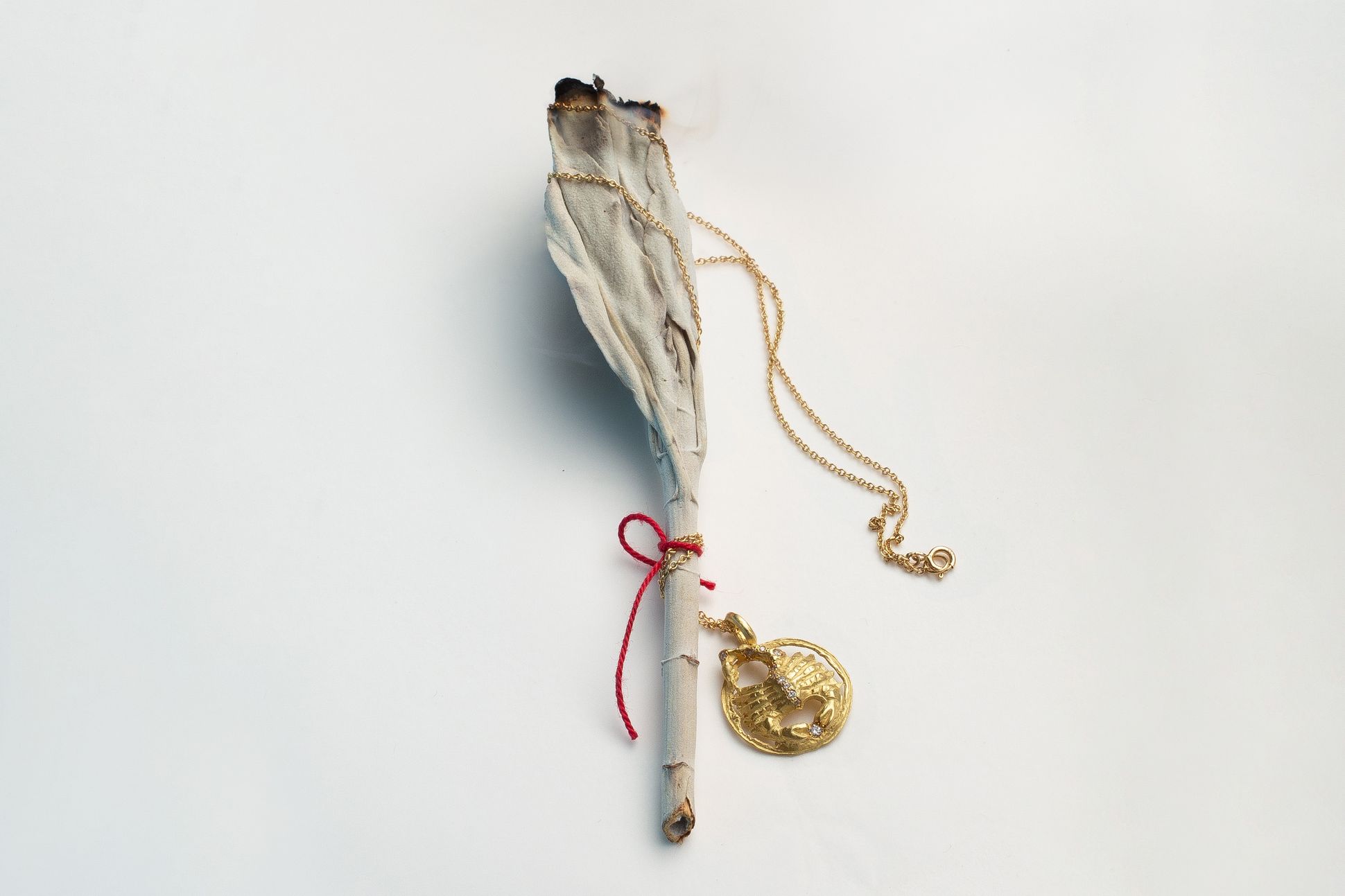 Collier en or avec pendentif rond sculpté, posé sur un bâton de sauge blanche partiellement brûlé et attaché par un fil rouge, sur fond blanc.