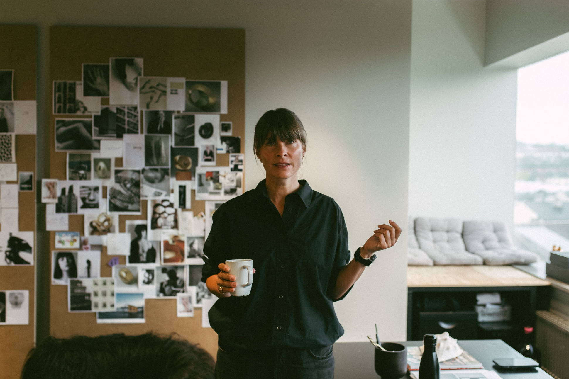 Une femme aux cheveux bruns avec une frange, vêtue d’une chemise noire, se tient debout dans un bureau lumineux. Elle tient une tasse blanche dans une main et semble parler. Derrière elle, un grand panneau recouvert de photos et d’images d’inspiration est accroché au mur. À droite, on aperçoit une banquette avec des coussins gris clair et une grande fenêtre donnant sur une vue urbaine.