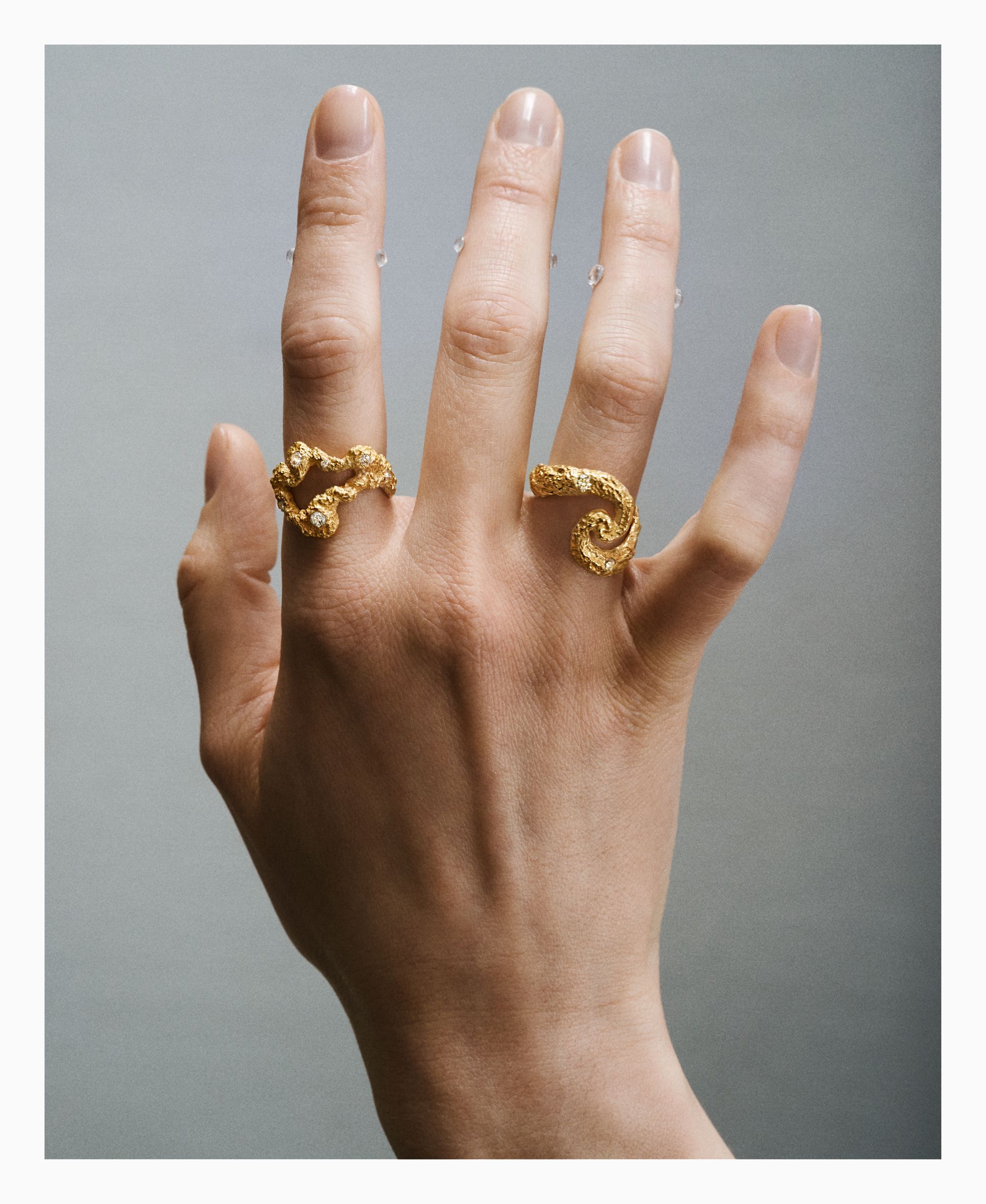 Hand wearing two sculptural gold rings by Griegst, with organic, textured forms and small diamonds, photographed against a soft grey background.