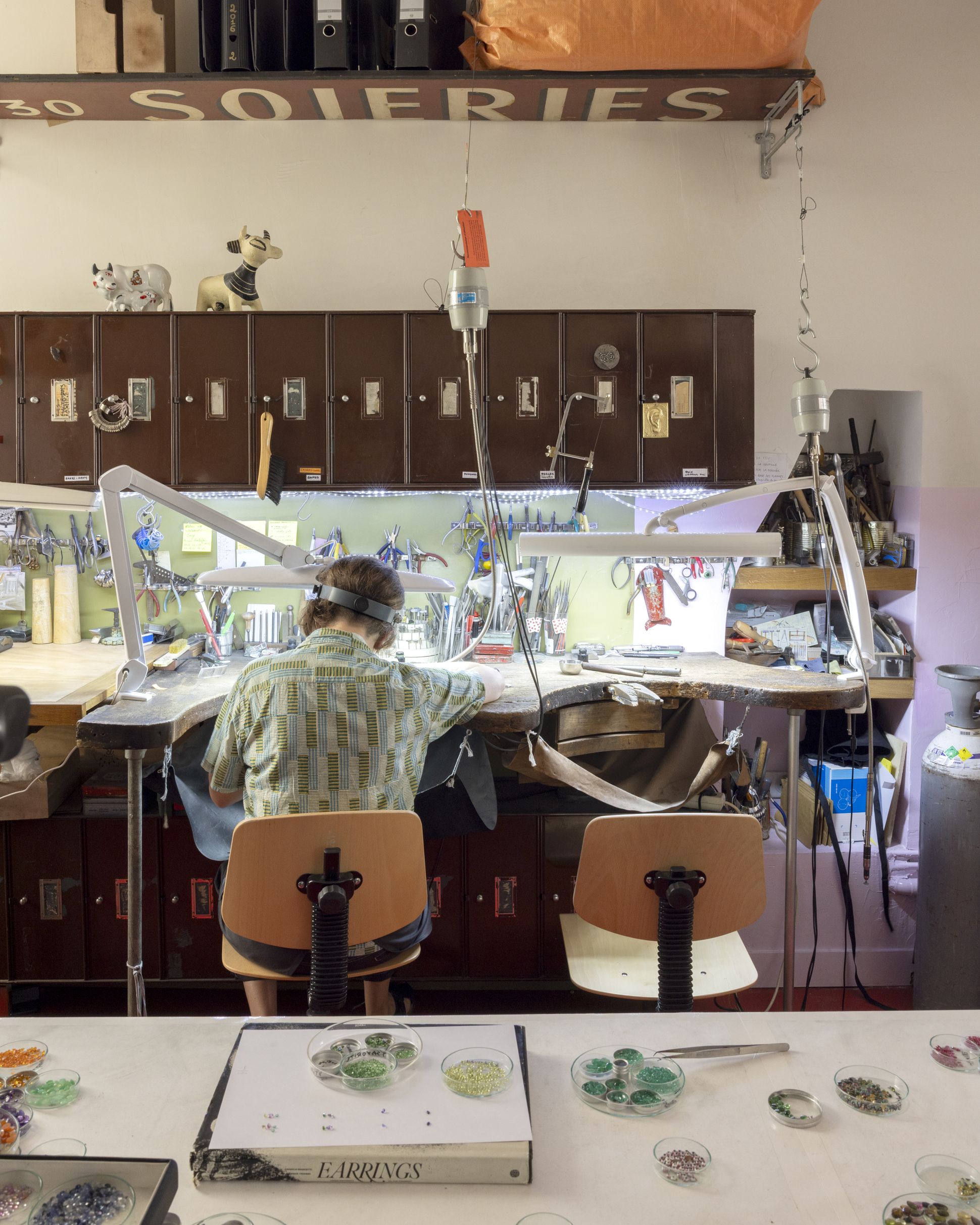 Jeweller at a workbench in the Dorette atelier, surrounded by tools, lamps and trays of colourful gemstones, seen from behind during the crafting process.