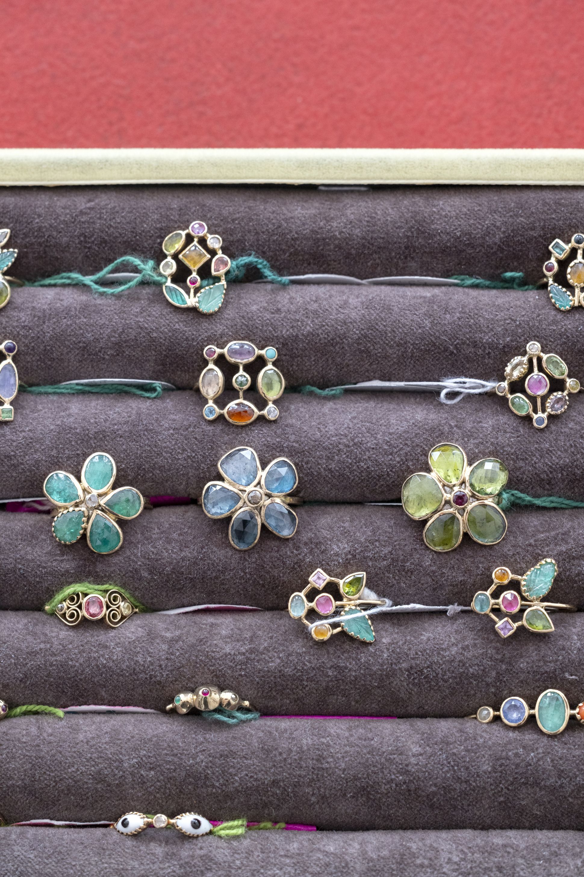 Assortment of Dorette rings with colourful gemstones in floral and geometric settings, displayed in a velvet tray against a red background.