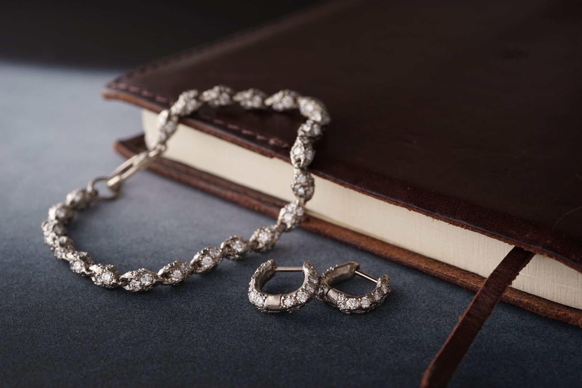 Bracelet et boucles d’oreilles en or blanc sertis de diamants, posés sur un carnet en cuir brun.