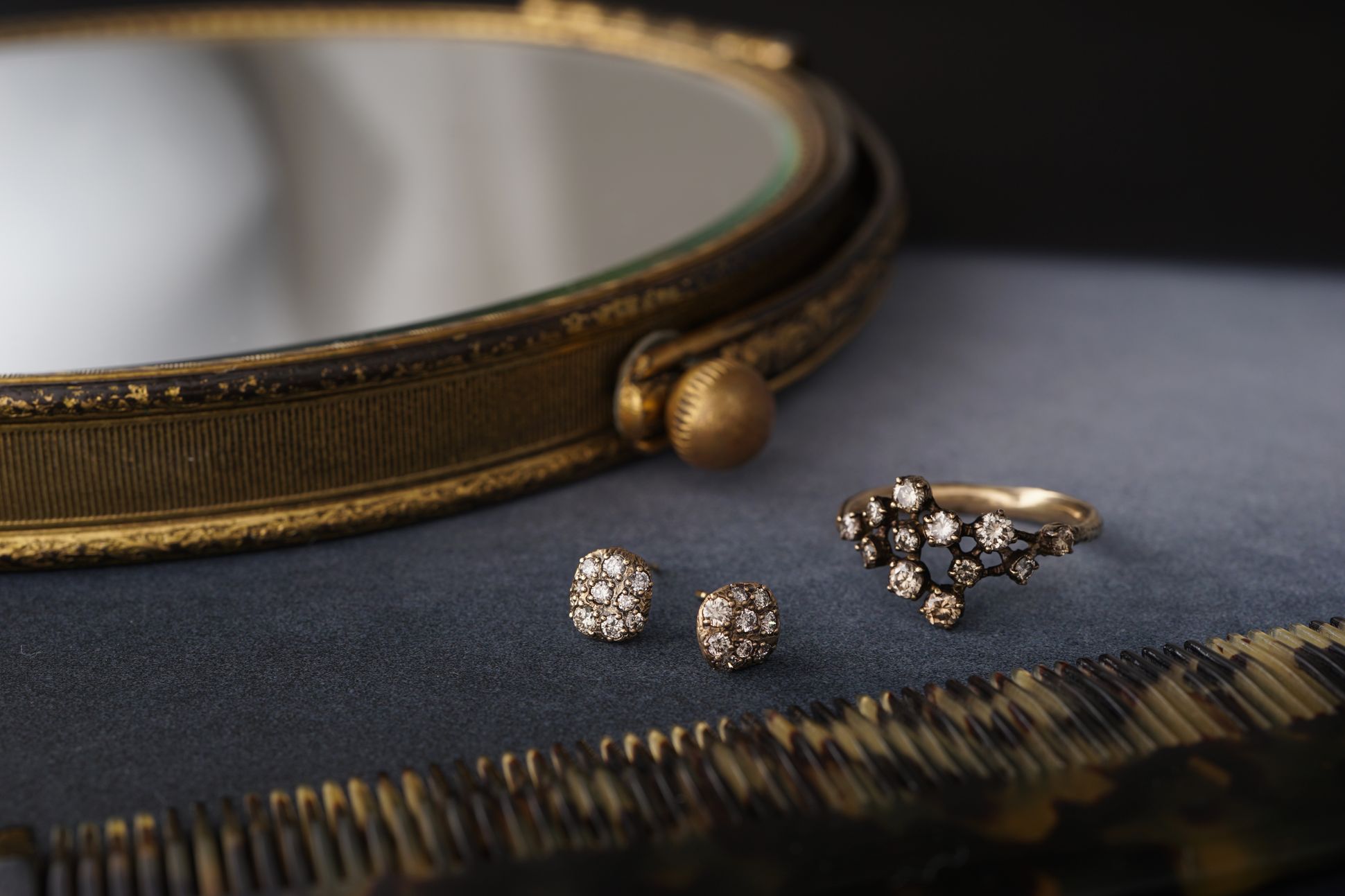 Bague et boucles d’oreilles en or serties de diamants, présentées sur une surface sombre à côté d’un miroir ancien et d’un peigne en écaille.