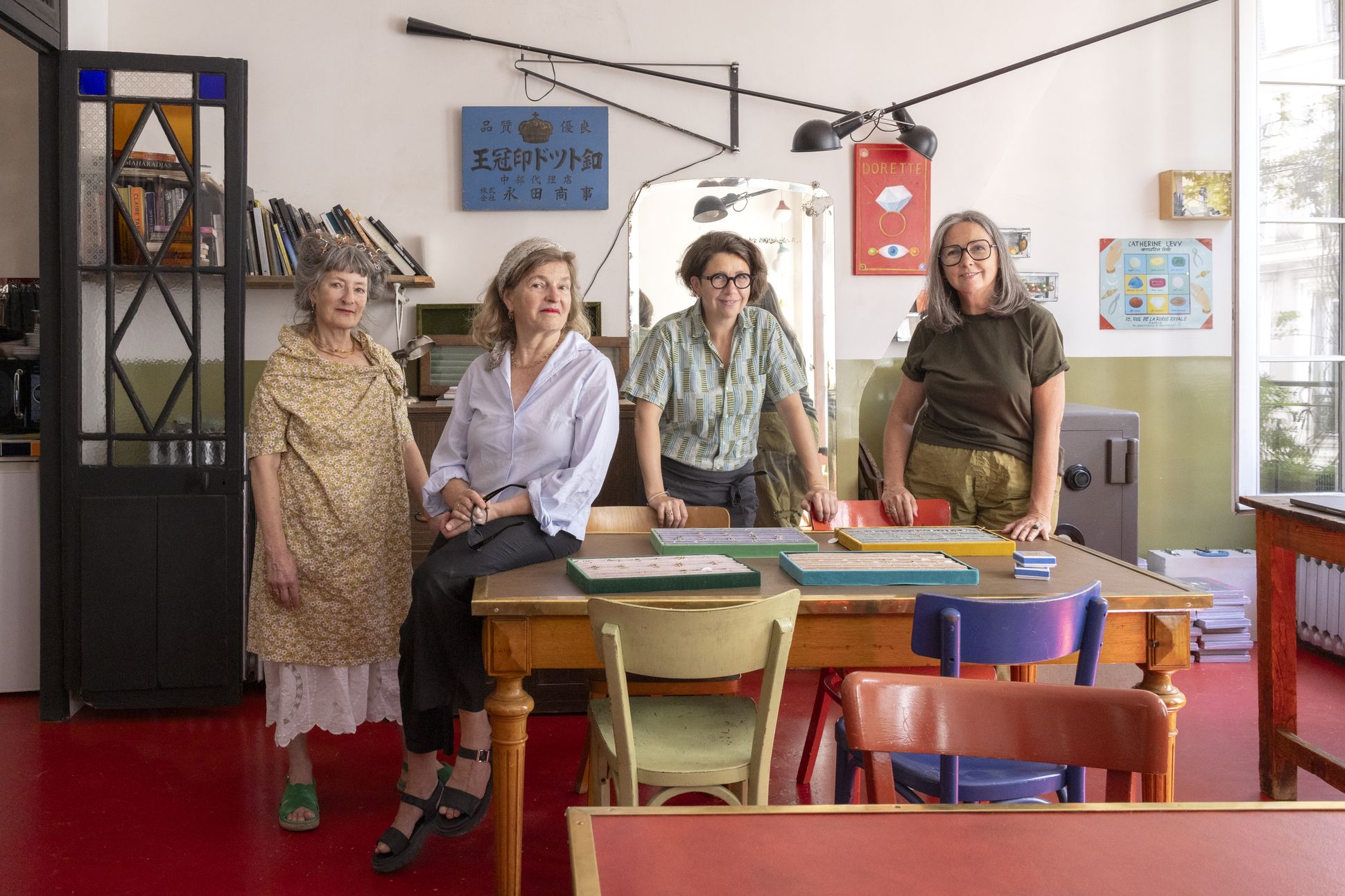 Four women standing around a wooden table displaying trays of Dorette jewellery, inside a colourful, eclectic studio with bookshelves, art prints and natural light from large windows.