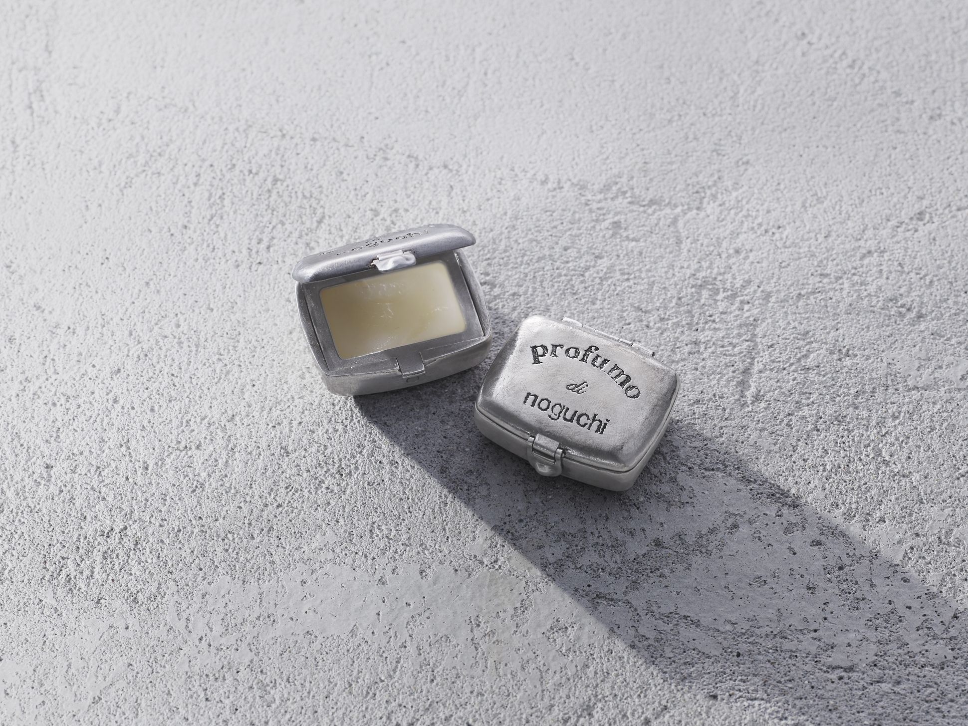 Parfum solide “Profumo di Noguchi” présenté dans un écrin métallique gravé, posé sur une surface minérale grise.