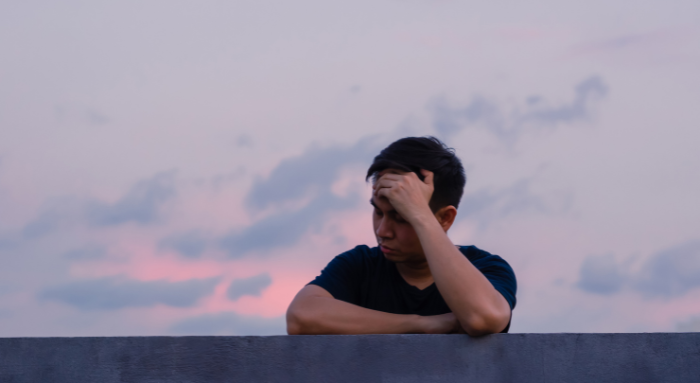 A man looks worried and deep in thought leaning on a wall against a pink sunset sky