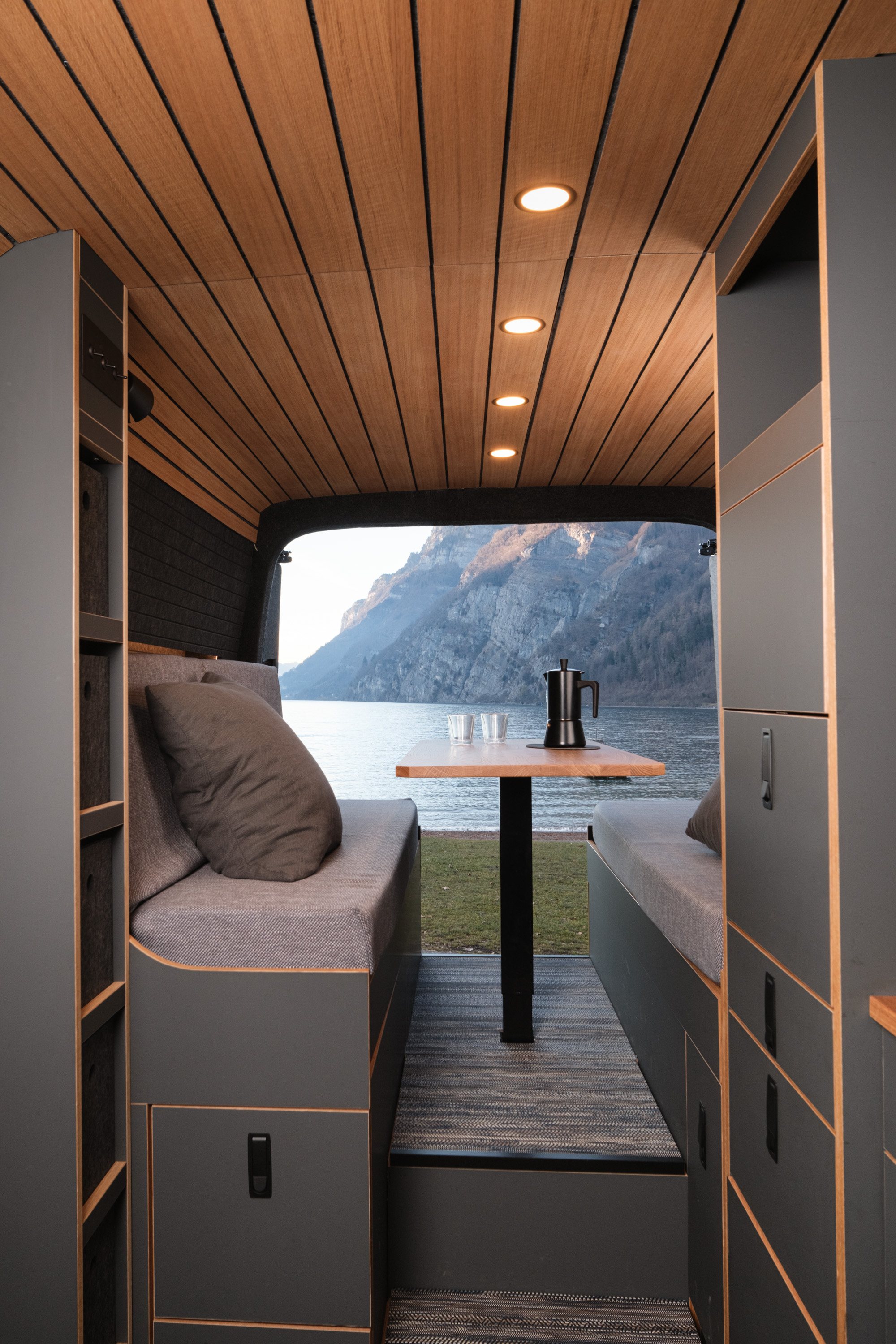 Blick von vorne durch den Camper Van Patgific von Cocoon Vans. Die Hecktüren sind geöffnet und geben den Blick auf einen See und Felswände die bis ins Wasser reichen frei. Die Beleuchtung in der Eichendecke ist eingeschaltet. Das aufahrbare Bett ist zu einer Sitzlounge umfunktioniert. Auf dem Tisch steht eine Kaffeekanne und zwei Tassen