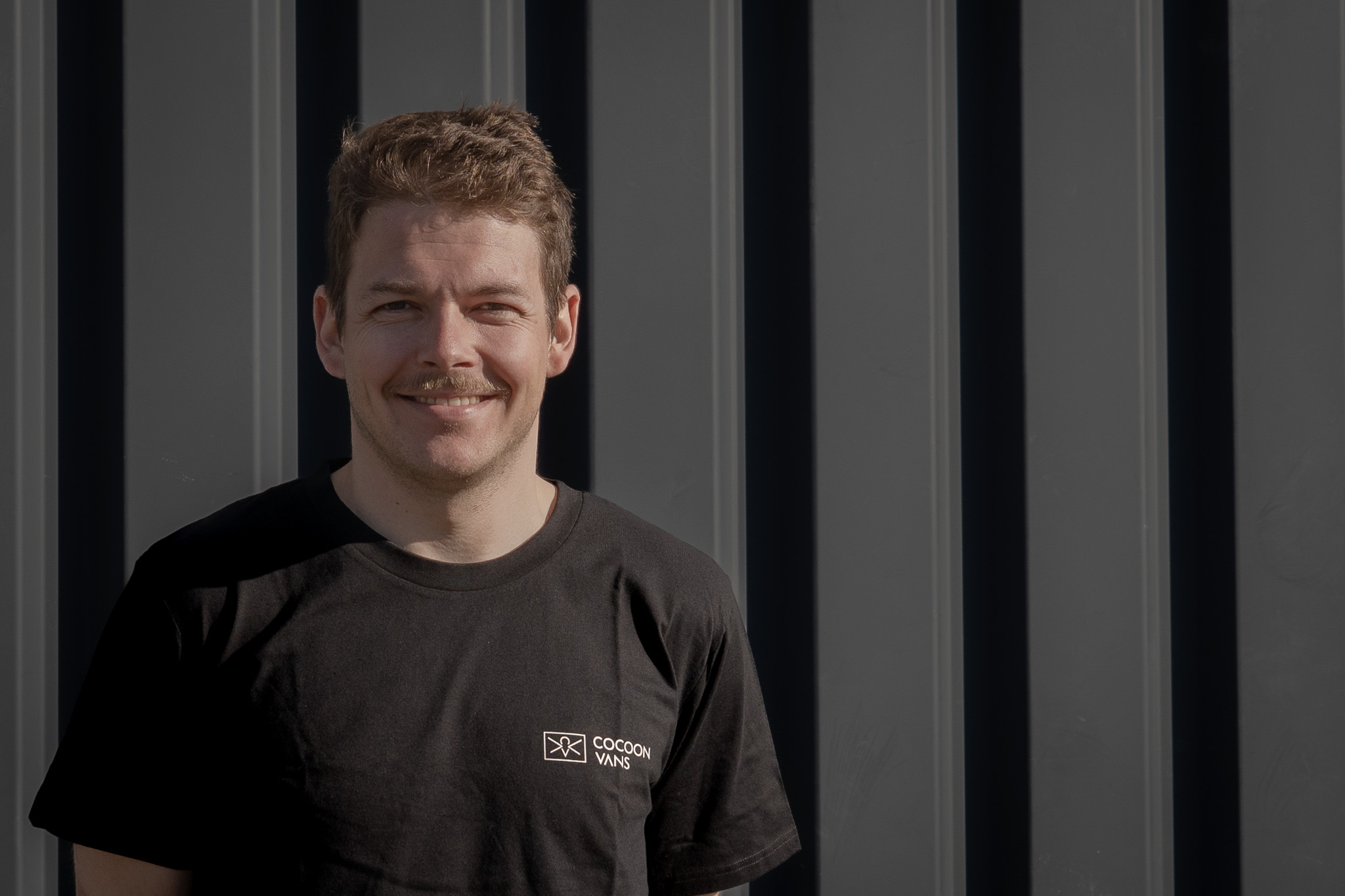 Jonas trägt ein dunkles T-Shirt mit dem Cocoon Vans Logo und lächelt in die Kamera. Er steht vor einer dunklen Wand im freien