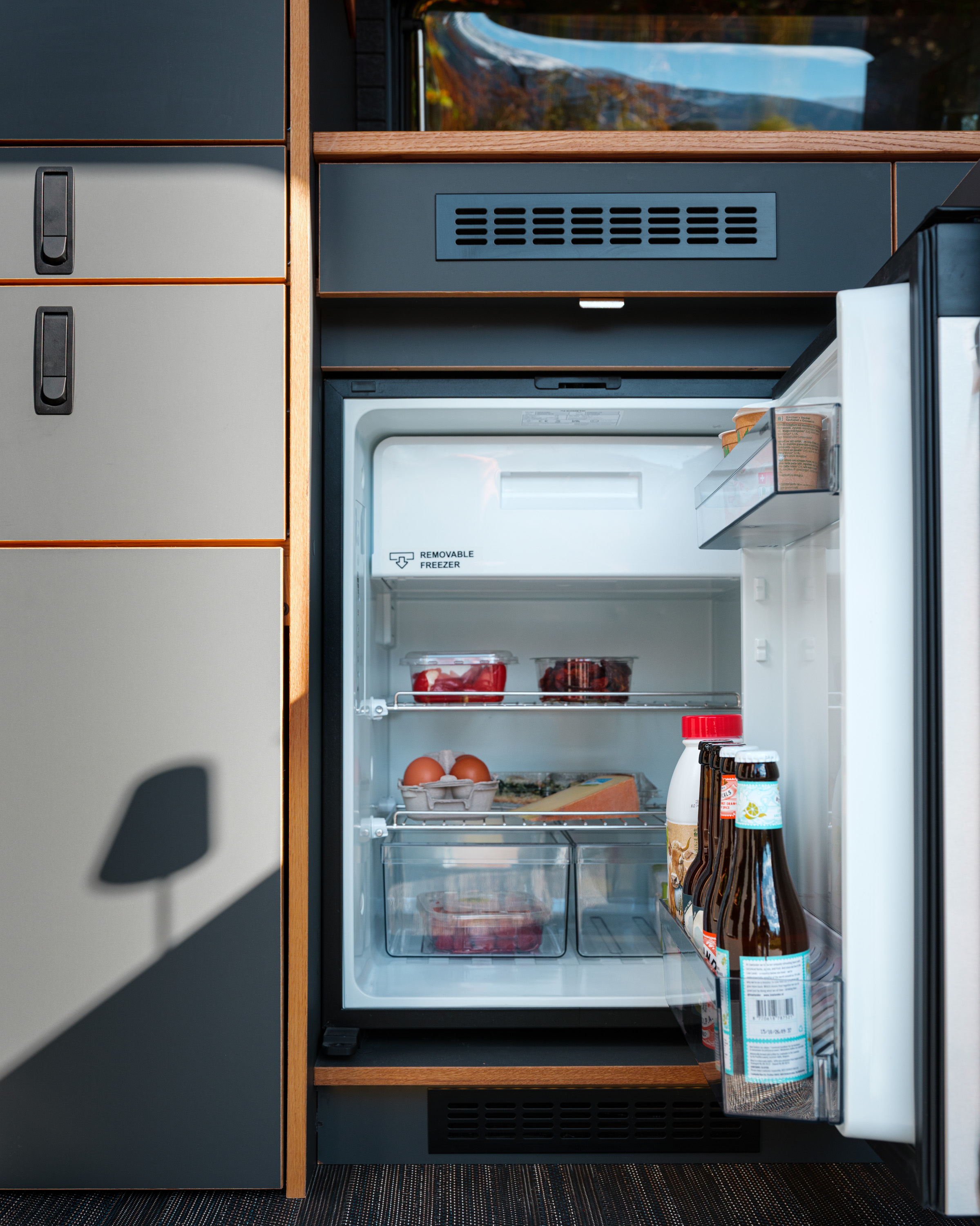 geöffneter Kühlschrank im VW Crafter Patgific von Cocoon Vans. Die fronten sind antrazit die Kanten aus Eiche. Der Kühlschrank ist geöffnet und gefüllt mit Getränken, Früchte, Käse und Eier