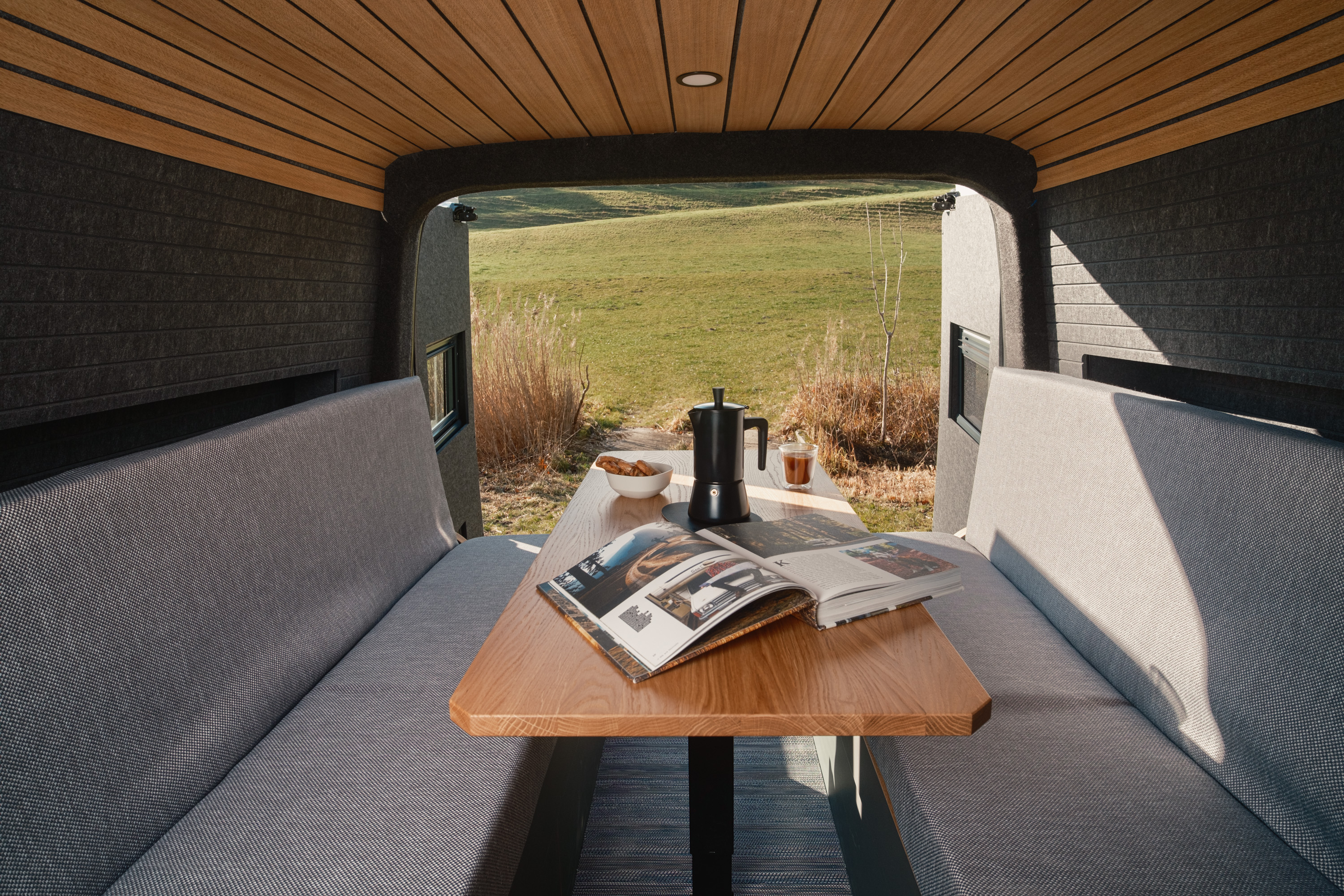 Kleinserie Patgific von Cocoon Vans.Graue Sitzbank vor Anthrazitfarbener Seitenwand. Elektrischer Hubtisch mit Kaffeekanne und Reisebuch. Hecktüre geöffnet mit Sicht auf die grüne Landschaft