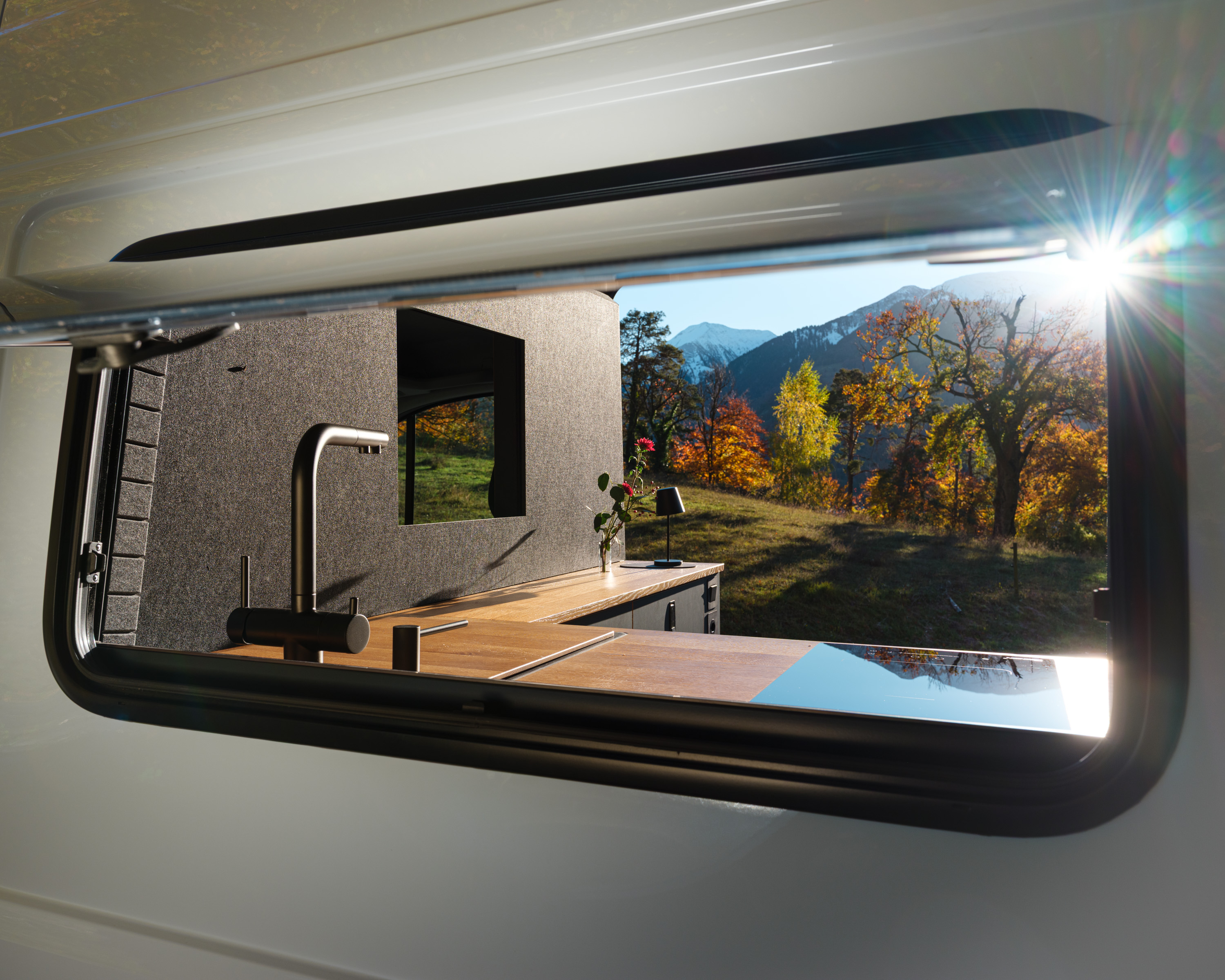 Blick durch das Seitenfenster des VW Crafters Patgifif. Im Camper Van sieht man die Abdeckungen der Küche aus Eiche, das Induktionskochfeld, den schwarzen Wasserhahn. Die Schiebetüre ist geöffnet und der Blick geht raus in eine Herbstlandschaft mit farbigen Blättern, weissen Bergen und die Sonne die gleich hinter den Gipfeln verschwindet