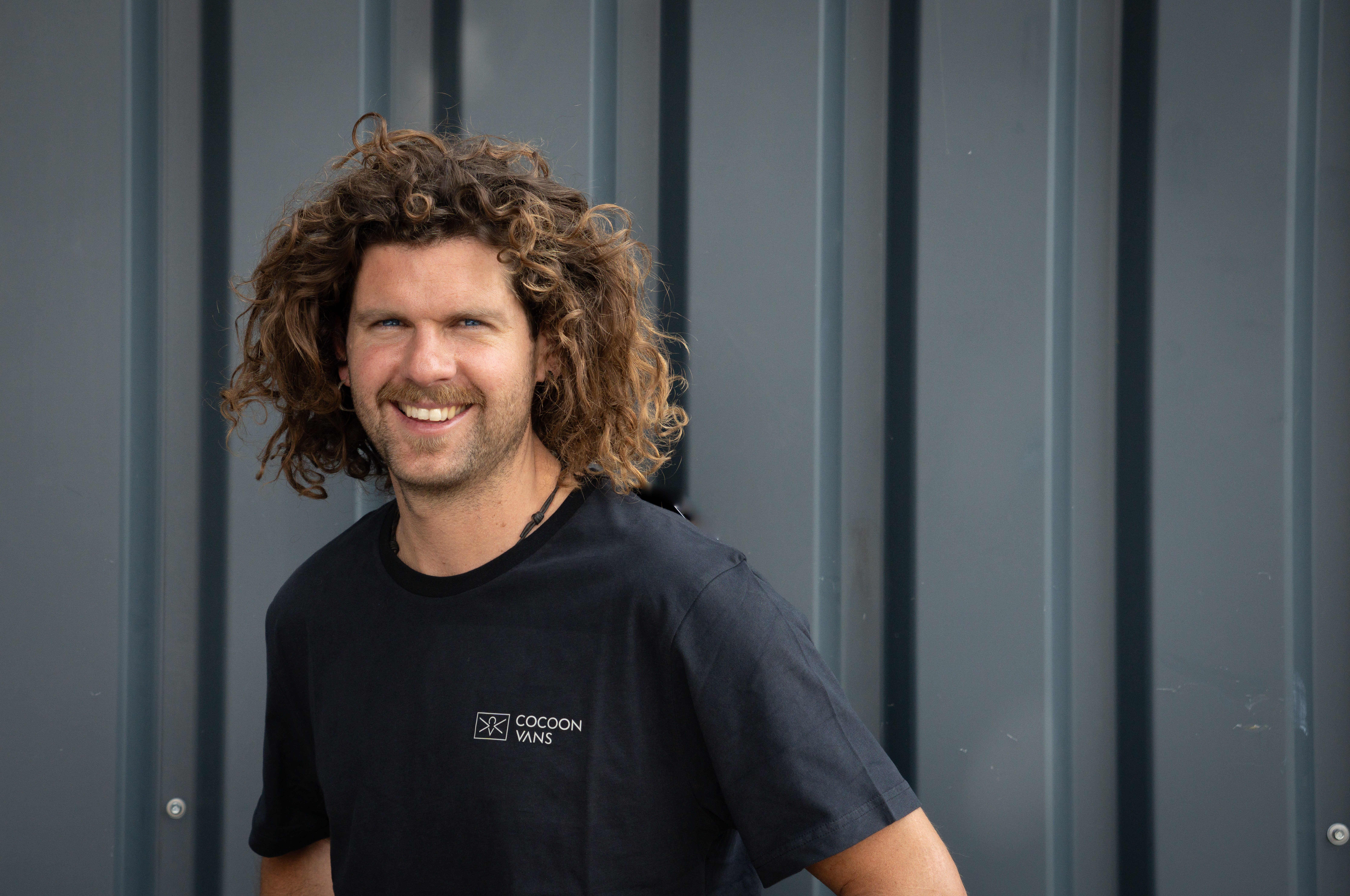 Adi steht im freien und hat im rechten Ohr ein grösseres Ohring. Der Hintergrund ist aus dunklem Wellblech