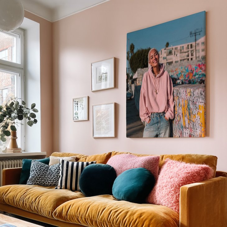 Großer Leinwanddruck mit Porträtmotiv an einer rosa Wand über einem senfgelben Samtsofa mit bunten Kissen