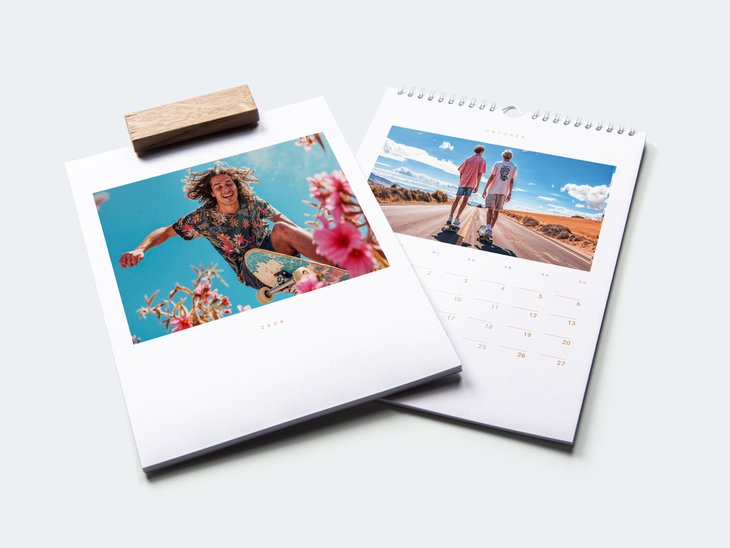 Deux calendriers muraux sont superposés et montrent la couverture ainsi qu'une page du calendrier avec des portraits. L'un des calendriers est muni d'un élégant crochet en bois, l'autre d'une reliure spirale blanche.