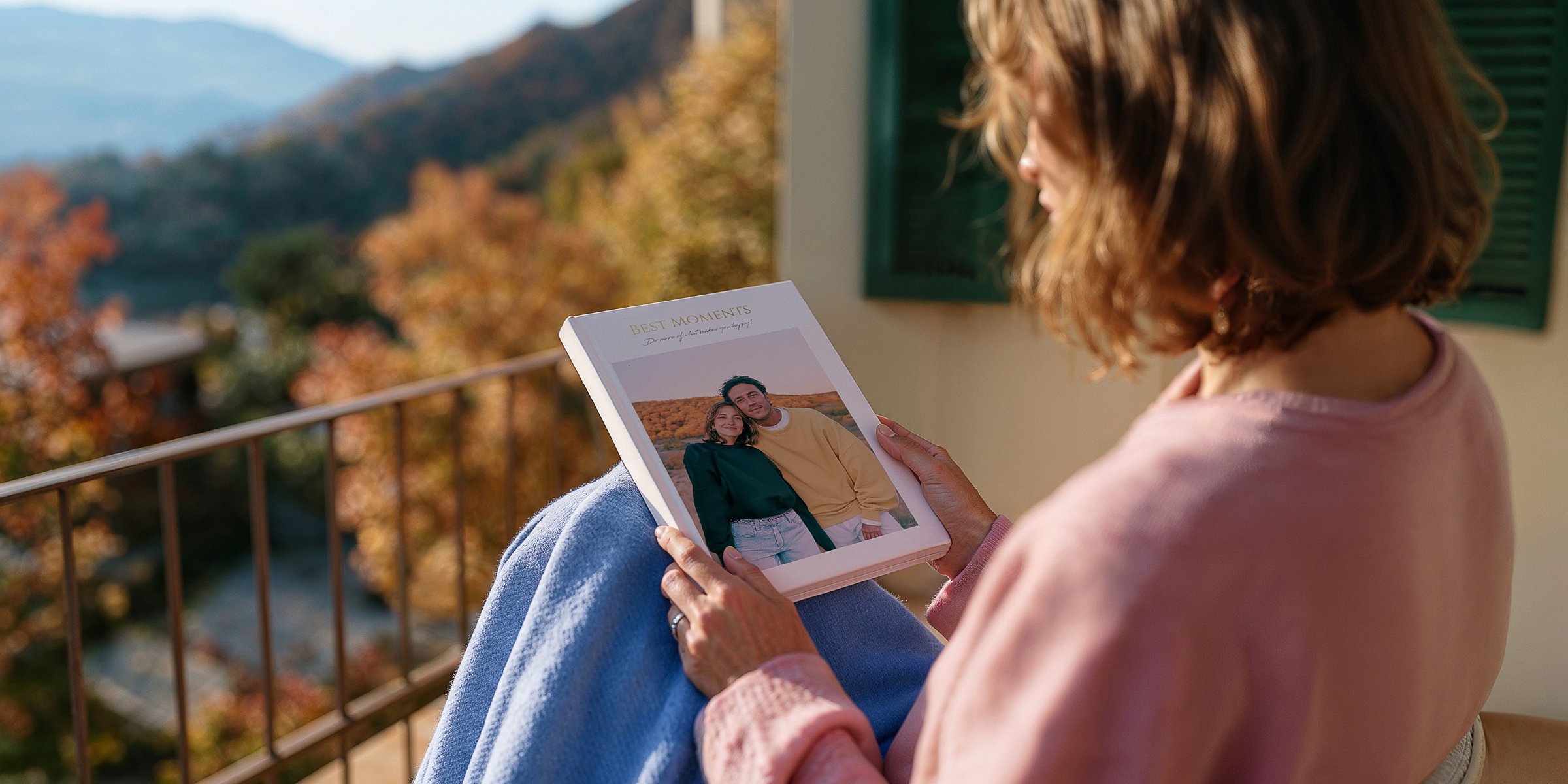 Frau sitzt auf einem Balkon mit Herbstlandschaft und hält ein Fotobuch mit dem Titel 'Best Moments' und einem Paar-Foto auf dem Cover