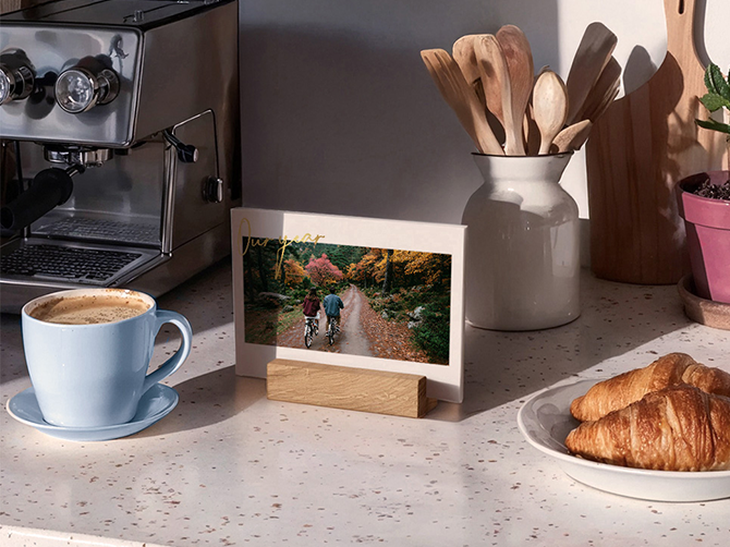 Tischkalender im Querformat mit Holzfuß und Herbstfoto zweier Radfahrer neben Kaffeetasse, Espressomaschine und Croissants