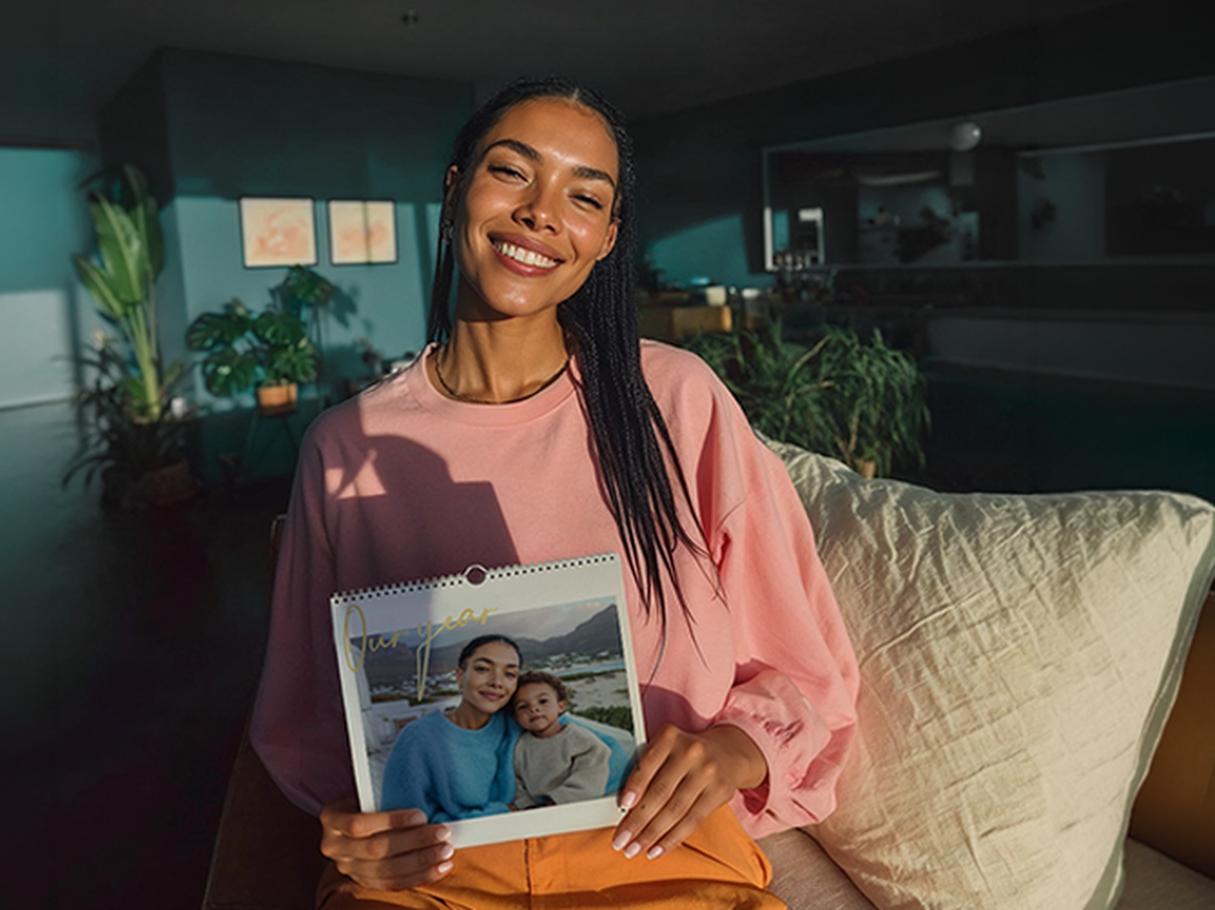 Lächelnde Frau mit Zöpfen sitzt auf einem Sofa und hält einen Querformat-Wandkalender mit Foto von Mutter und Kind