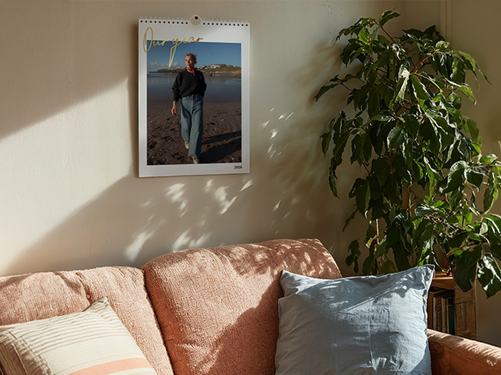 Hochformatiger Wandkalender mit Strandfoto hängt an heller Wand über einem lachsfarbenen Sofa neben einer Zimmerpflanze