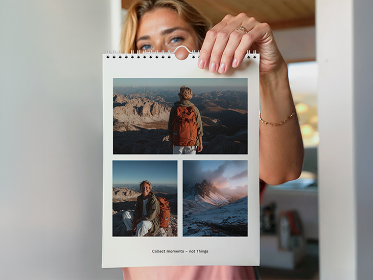 Frau hält einen Hochformat-Wandkalender mit drei Bergfotos einer Wanderin mit rotem Rucksack in der Hand