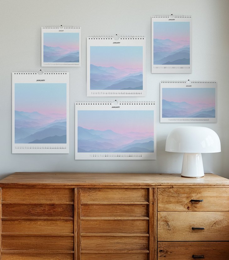 Sechs Wandkalender in verschiedenen Größen und Formaten mit Bergmotiv hängen über einer Holzkommode mit weißer Lampe