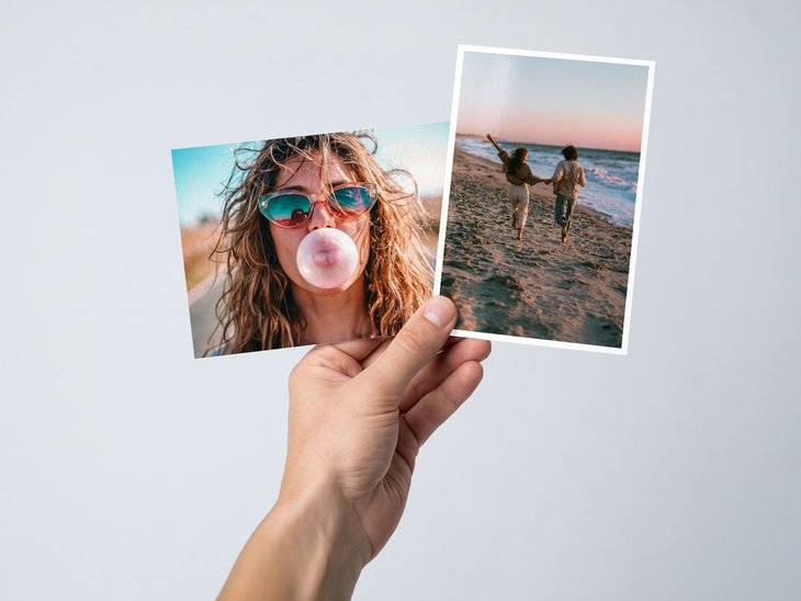 Hand hält zwei Fotoabzüge im Format 10 × 15 mit Urlaubs-Selfie und Strandpaar-Motiv vor hellem Hintergrund