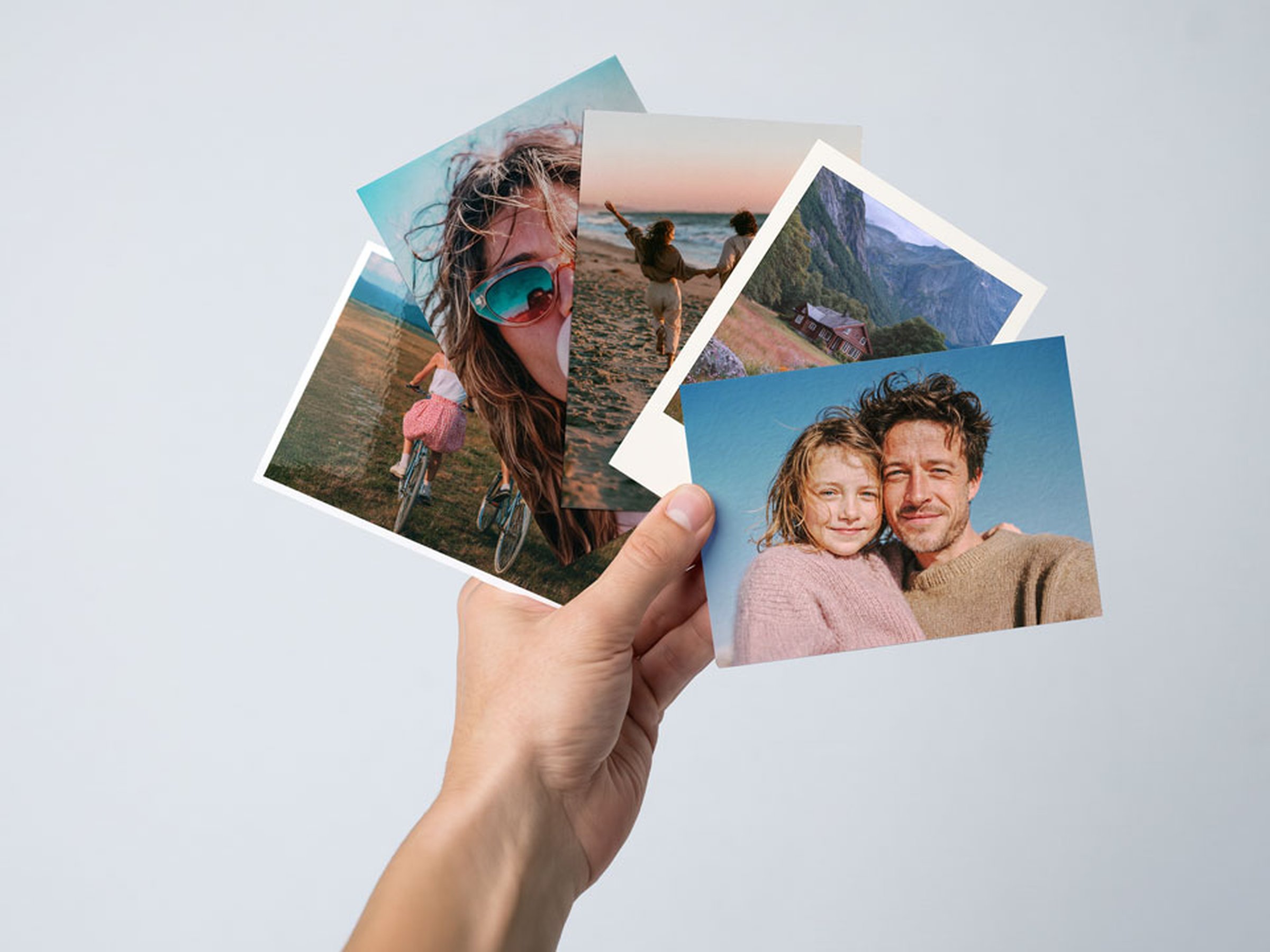 Hand hält vier aufgefächerte Fotoabzüge mit Urlaubs- und Familienmotiven vor einem hellen Hintergrund