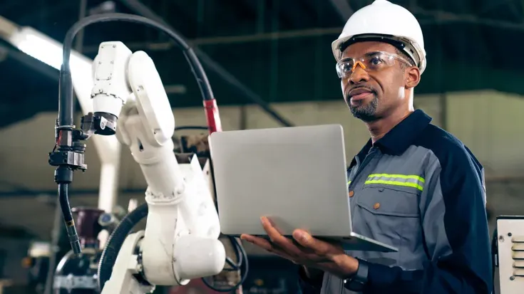 Trabalhador com capacete e óculos de proteção usando notebook ao lado de um cobot, ilustrando a colaboração entre humanos e máquinas na indústria 5.0