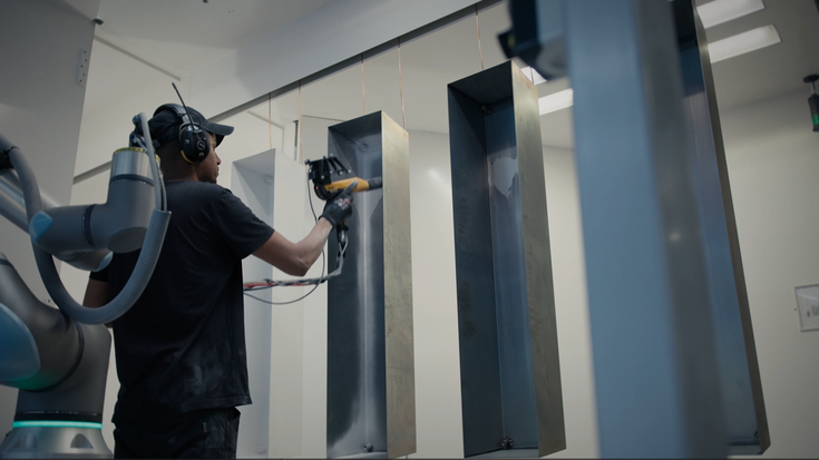 Industrial collaborative robot assisting with spray painting of tall metal panels in a clean, enclosed industrial workspace, while a technician wearing protective gear guides the process.
