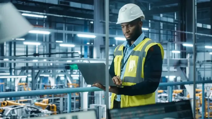 Engenheiro analisa dados em laptop dentro de uma fábrica automatizada, representando estudos de capacidade ociosa na produção industrial.