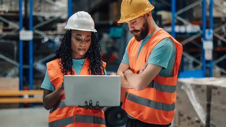 Dois trabalhadores num armazém consultando um laptop com um sistema MES em uso para controlar a produção.