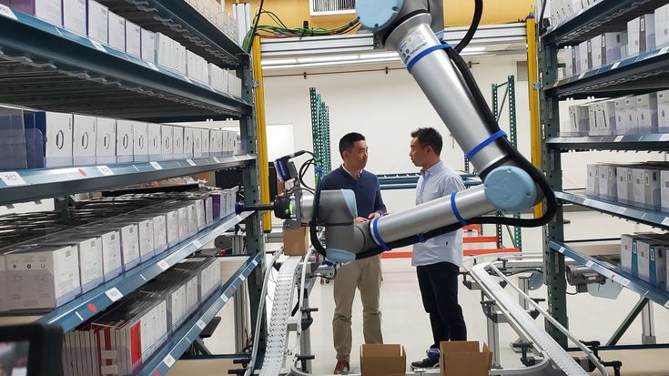 Universal Robots collaborative robot (cobot) automating warehouse order fulfillment, picking and placing boxed products on shelving with integrated conveyor system, showcasing flexible logistics and intralogistics automation for e-commerce and distribution centers