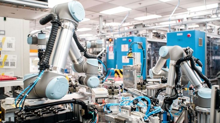 Universal Robots cobot arms operating on an automated assembly line in a factory, surrounded by tooling and cables.